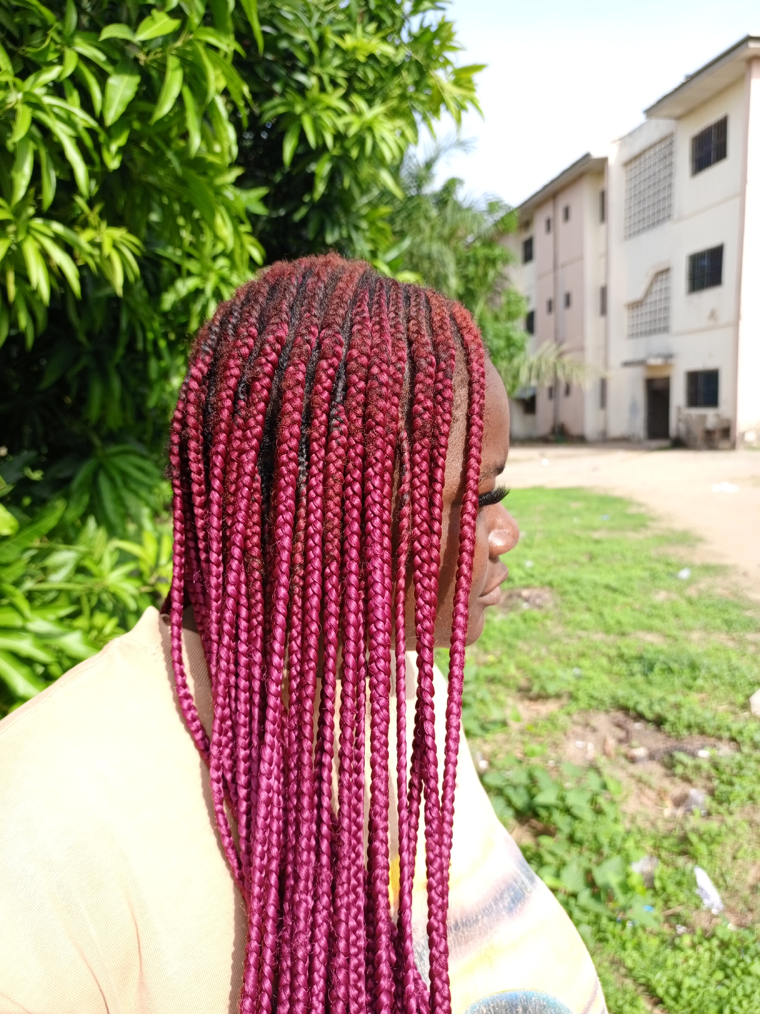An image of a woman with maroon box braids outdoors in Keffi, Nigeria. The woman is in profile, looking right. Her skin is brown, and she is wearing false eyelashes. She is wearing a light-colored shirt. The background includes green foliage, a beige building, and a grassy area with a dirt path.