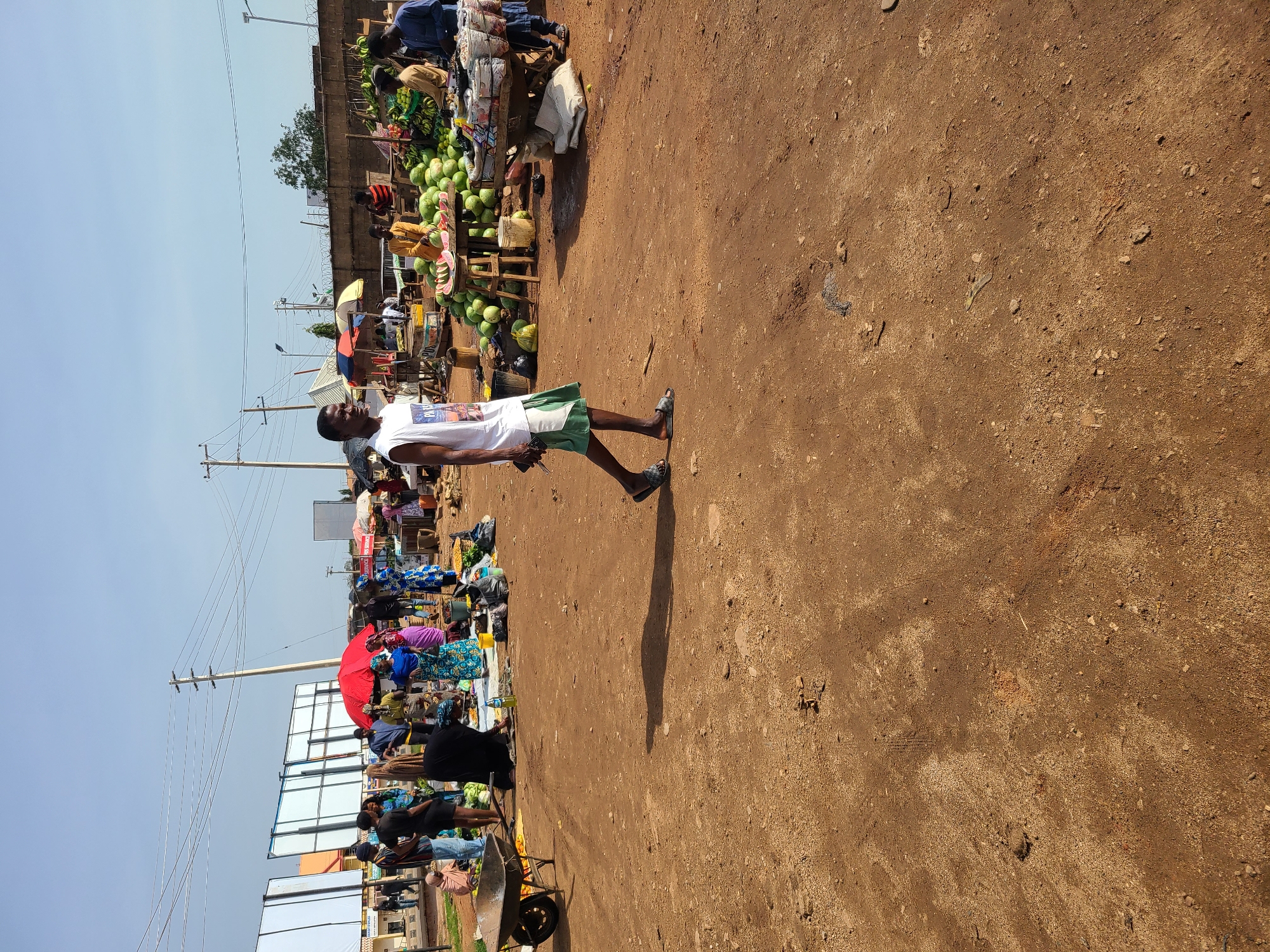 The image depicts an outdoor market scene in Gura Top, Nigeria, on a clear, sunny day. The ground is covered in reddish-brown dirt. In the foreground, a young man walks from right to left, wearing a white sleeveless t-shirt with a graphic print and green shorts. He is looking to his left. Behind him, a variety of market stalls are visible. To the left, a wheelbarrow is parked on the dirt. Further back, several people are gathered around stalls displaying produce, including a large pile of watermelons. Some individuals are wearing colorful clothing. In the background, power lines are strung between utility poles, and billboards are positioned along the roadside. The overall impression is of a busy, open-air marketplace.