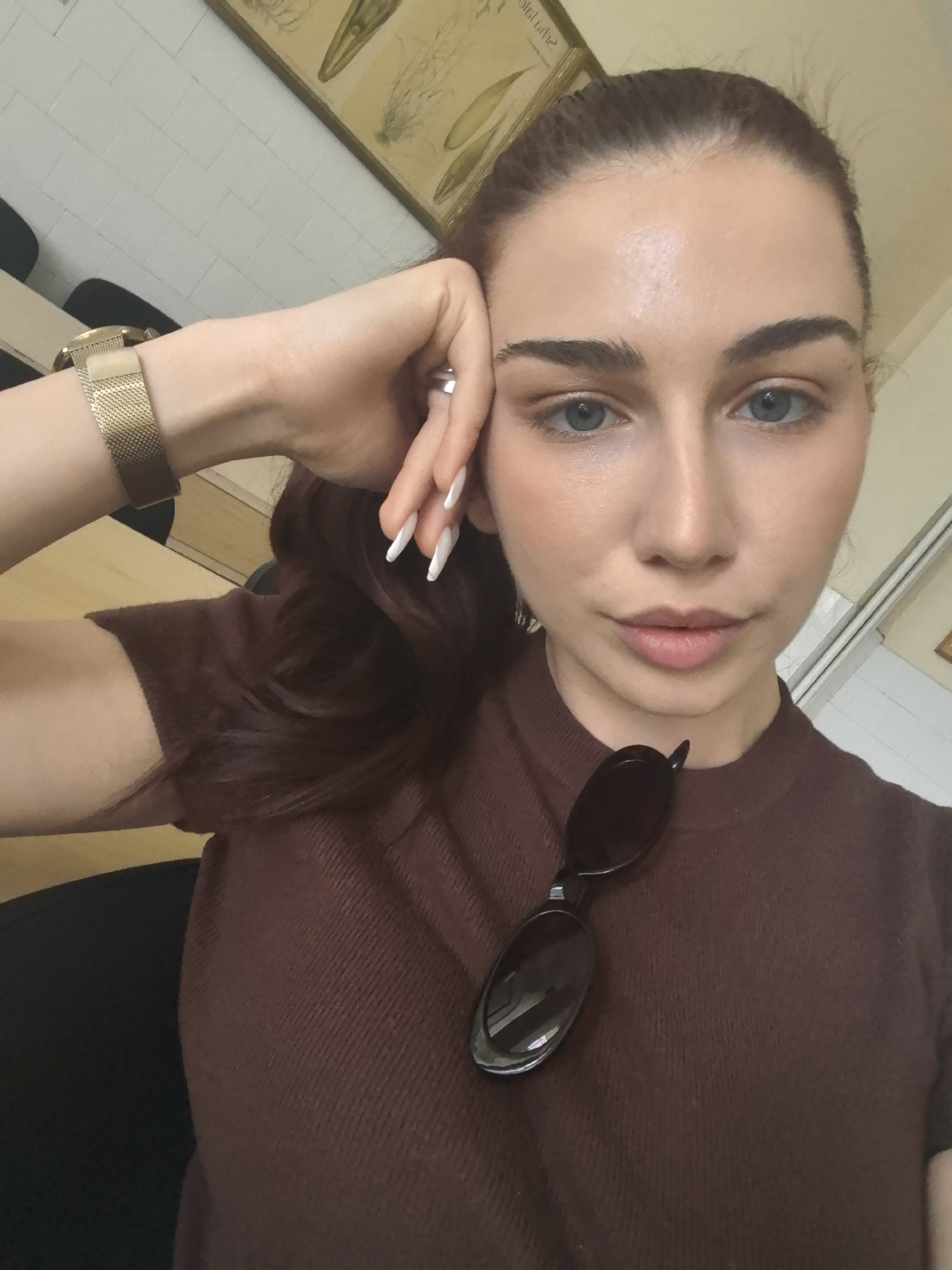 A young woman with fair skin, blue eyes, and dark brown hair pulled back into a ponytail is pictured in a close-up, selfie-style shot. She is wearing a brown, ribbed, short-sleeved shirt, and has a pair of dark sunglasses resting on her chest. Her left hand is resting on her forehead, with her fingers extended, and she is wearing a gold watch on her left wrist and a ring on her left index finger. The background appears to be an indoor setting with white tiled walls and some framed artwork on the upper wall. There is also a glimpse of wooden furniture and a black chair in the background. The image is well-lit, with the woman's face being the primary focus. There are no specific indicators in the image to suggest a location within the city of Bogomilovo, Bulgaria.