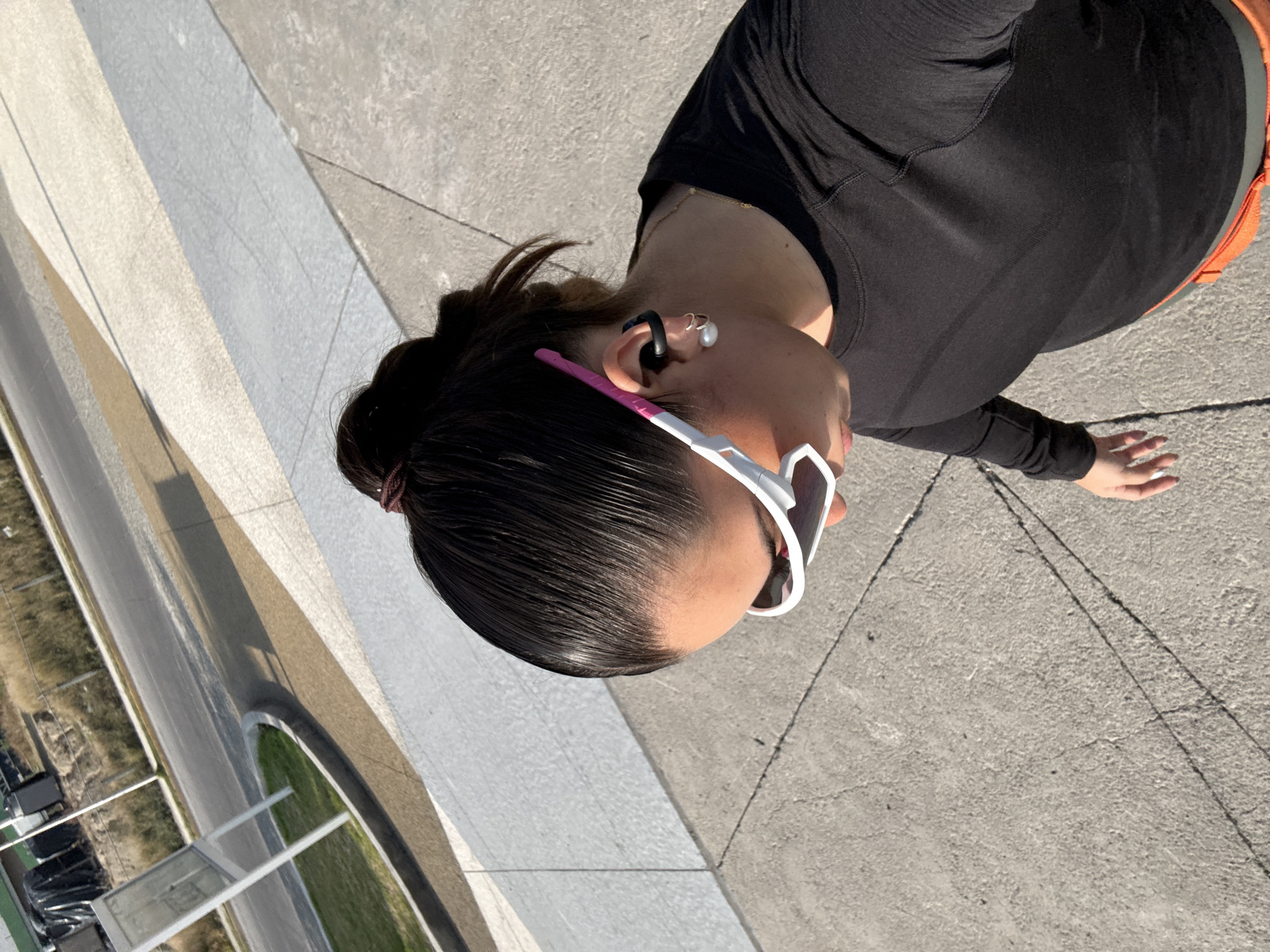 A high-angle shot shows a woman standing on a concrete surface. She has dark hair pulled back in a bun and is wearing white sunglasses with pink accents, a black long-sleeved shirt, and orange shorts. She has an earring and a wireless earbud in her left ear. To the left of the woman, a road and green space are visible. The setting is likely near or at the Museo Internacional del Barroco in Puebla, Mexico, considering the unique architecture and design of the surrounding area.