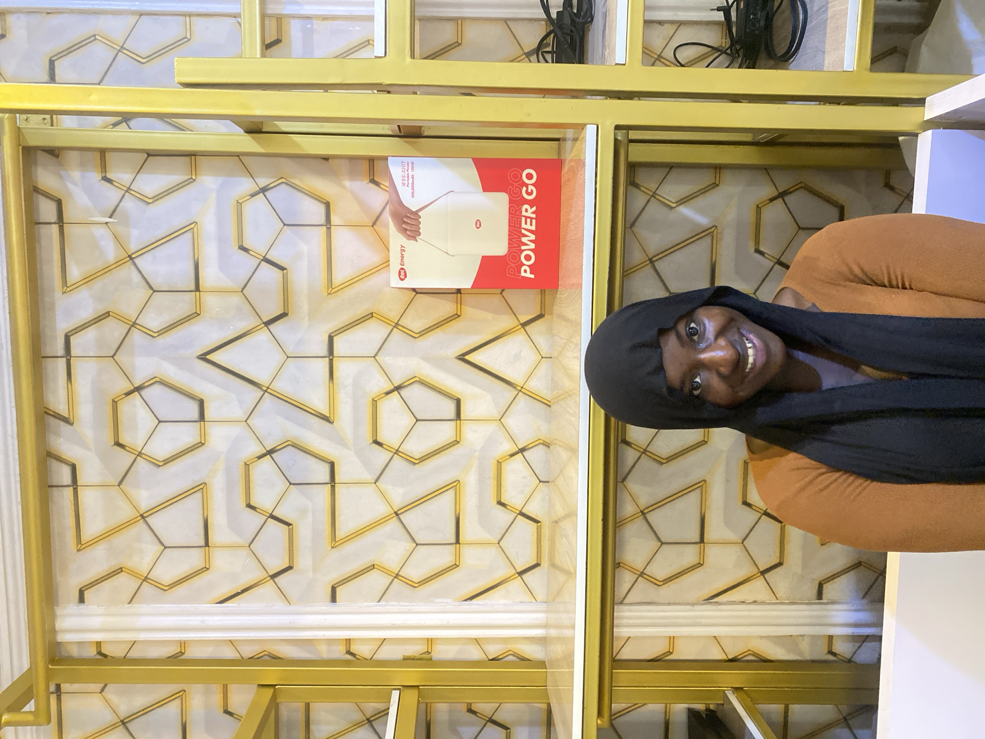 A young Black woman with a headscarf and an orange sweater smiles at the camera, positioned in front of a gold shelf unit and a wall with a geometric pattern. She is smiling widely, her head tilted slightly to the left. The shelf unit is made of gold-colored metal and has white shelves. A red and white box with an image of a portable power bank is placed on one of the shelves. The background wall features a repeating pattern of gold hexagons and lines against a white or light-colored surface. The image appears to be taken indoors, possibly in a retail or display setting.