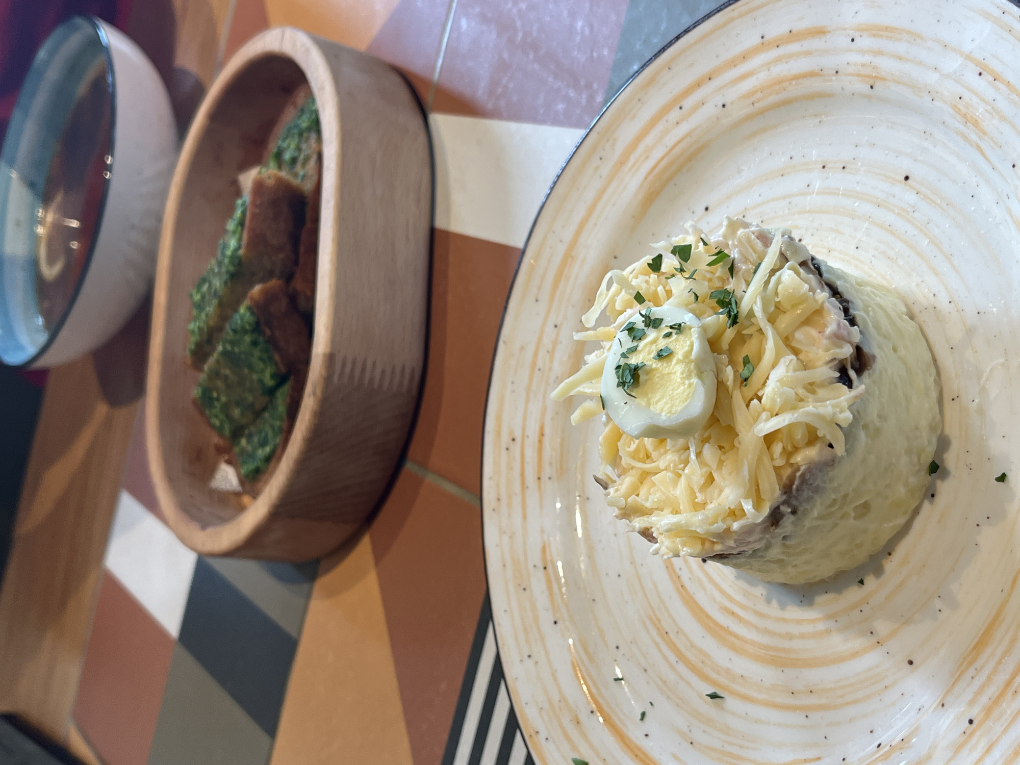 The image shows an overhead shot of a table setting with food items. A white ceramic bowl containing a dark liquid is on the left. Next to it is a wooden bowl filled with small, rectangular pieces coated in a green substance. A plate with beige swirls occupies the right side of the image. On the plate, a serving of food features what appears to be mashed potatoes, topped with shredded cheese, herbs, and half of a hard-boiled egg. The table surface has a geometric pattern with various colors including white, orange, brown, gray, and black. The location is Verkhnyaya Pyshma, Russia.