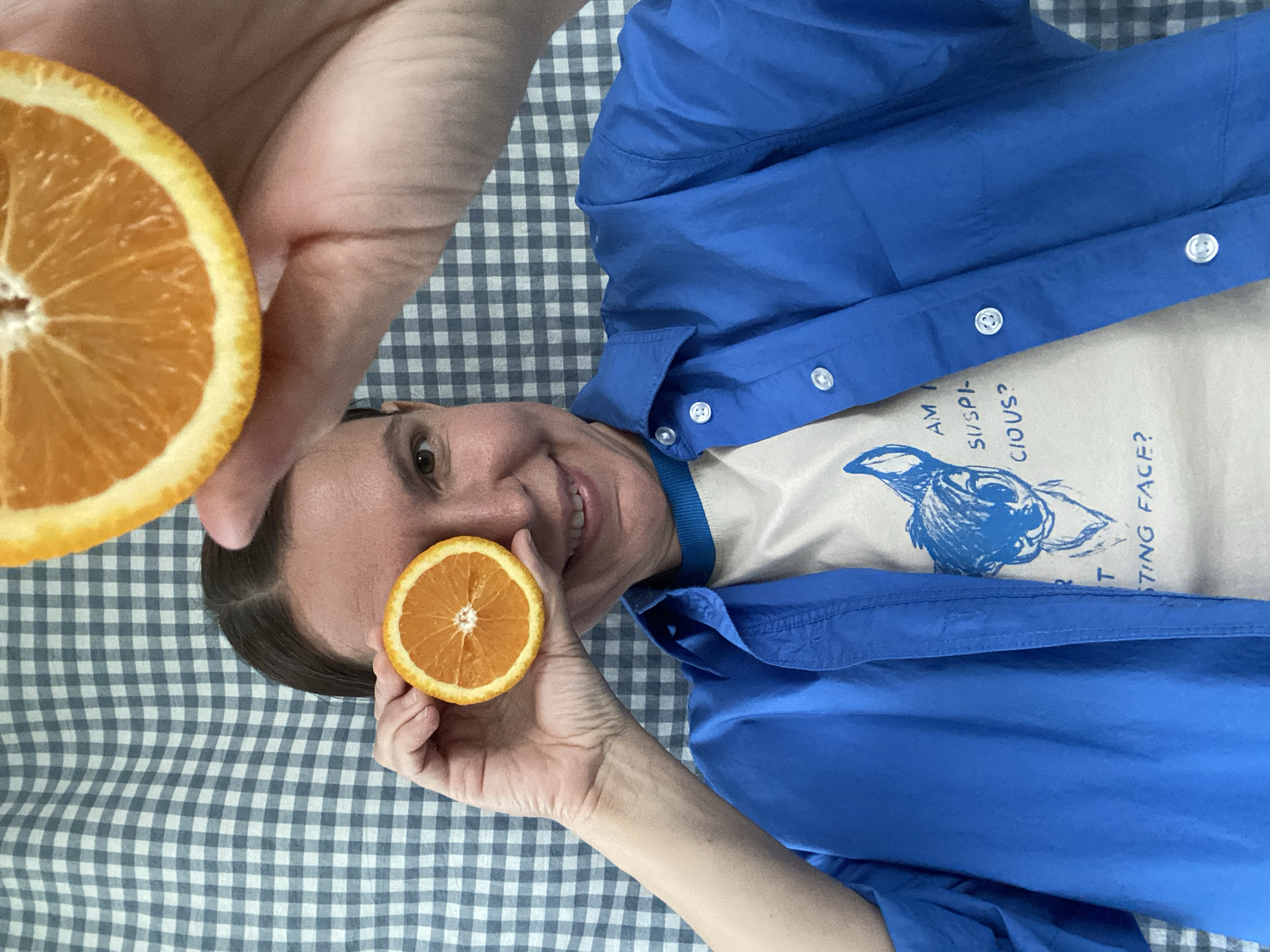 A person is laying on a light grey and white checkered surface, possibly a blanket or tablecloth. The person is smiling and has a slice of orange held up to their right eye. Another slice of orange is held in another person's hand, above the head. The person is wearing a light colored t-shirt with a blue graphic and text, and a blue, collared shirt. The background is out of focus, but appears to be an indoor setting. There is no indication that the image was taken in Chaikovsky, Russia.