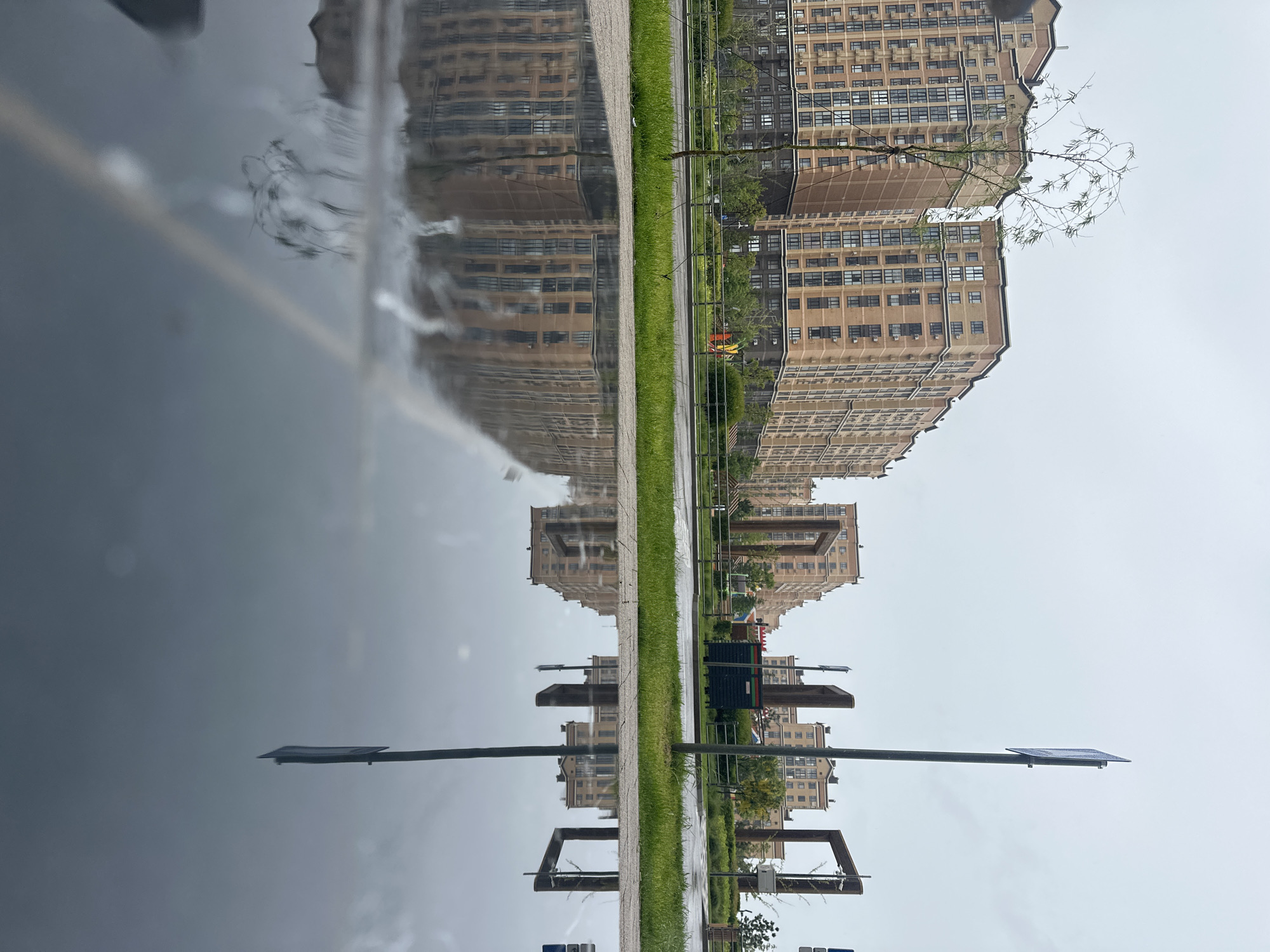 The image shows a wet pavement reflecting a street scene in what is purported to be Stavropol, Russia. The reflection includes a row of tall apartment buildings, a strip of green grass, and a walkway. Street lamps are visible in the foreground, partially submerged in the reflective surface. The sky is overcast. The water on the pavement creates a mirrored effect, blurring and distorting the reflected images. There is no indication of people or specific actions occurring in the scene.