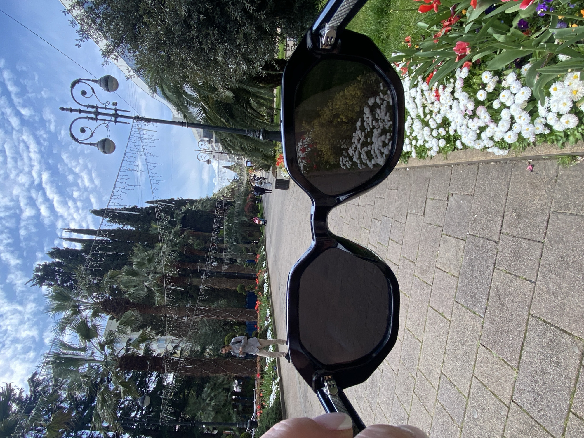 The image shows a person holding a pair of sunglasses in front of a scene in what appears to be a park or public space in Sochi, Russia.
The sunglasses are black and reflect the flowers and trees.
The background features a paved walkway, trees, including palm trees and pines, and flower beds filled with white and red flowers. There are decorative lamp posts and strung lights. A person can be seen walking on the walkway. The sky is blue with scattered clouds. The scene appears to be taken in a well-maintained public garden or park.