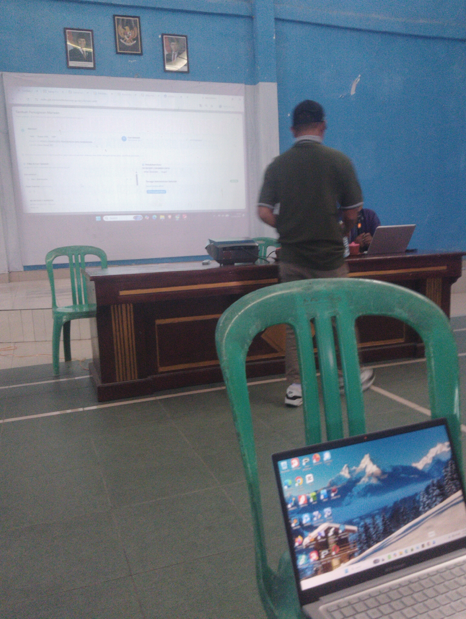 The image depicts a classroom or meeting room in Wanasaba, Indonesia. In the foreground, a green plastic chair is positioned in front of a laptop displaying a winter mountain scene as its wallpaper. The laptop is open and placed on a surface not fully visible. Behind this chair, a dark wooden desk with yellow accents is present. To the left of the desk, another green plastic chair stands empty.

Further back, a man wearing a dark green polo shirt and a dark baseball cap is standing, facing away from the camera, towards a large projection screen. The screen displays a webpage with text and icons, likely a presentation or online document. To the right of the standing man, a person in a purple shirt is seated behind the desk, with a laptop open in front of them.

The walls of the room are painted a light blue. Three framed pictures are hung on the wall above the projection screen. The floor is covered with dark grey tiling, marked with white lines, suggesting it might be a multi-purpose hall or a sports facility integrated with a learning space. The overall lighting suggests an indoor environment, possibly during daylight hours.