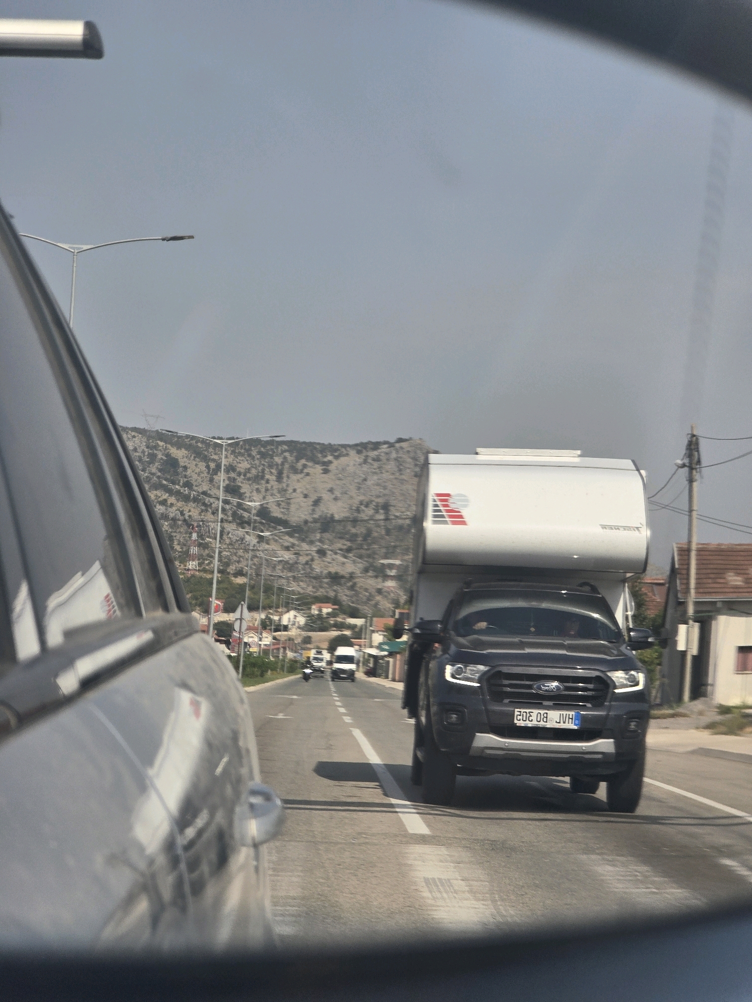 A dark Ford Ranger truck with a white camper shell is traveling on a highway, likely in Podgorica, Montenegro, given the license plate format. The truck has a license plate with the alphanumeric characters "20E 08 JVH". The truck is being viewed from the perspective of another vehicle, as indicated by the reflection of the truck and surrounding environment on the left-side car's window. In the background, there are hills with sparse vegetation, a few buildings, streetlights, and other vehicles on the road, including a white van and a motorcycle. The sky is clear and hazy.