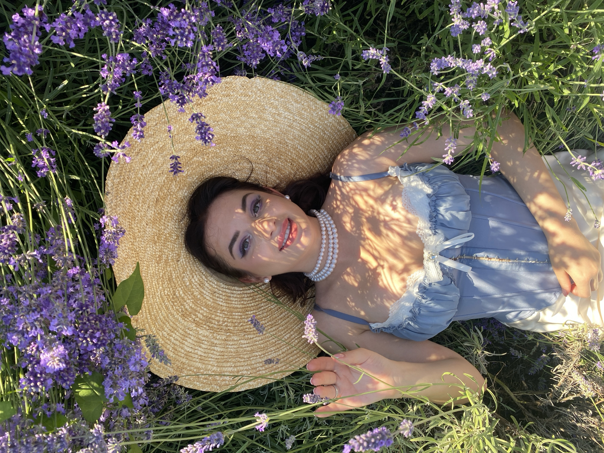 A woman is lying in a lavender field, her head tilted back and resting on a straw hat. She is wearing a light blue corset with white lace trim, a pearl necklace, and white skirt. Her eyes are closed, and her lips are slightly parted as she smiles. Her hand is raised to her face, and she is touching a lavender sprig. The lavender is in bloom, with purple flowers and green stems surrounding her. The sun is shining, casting dappled shadows on her face and chest. The image appears to have been taken from directly above, looking down at the woman in the field. The location is Verkhneye Uchdere, Russia.