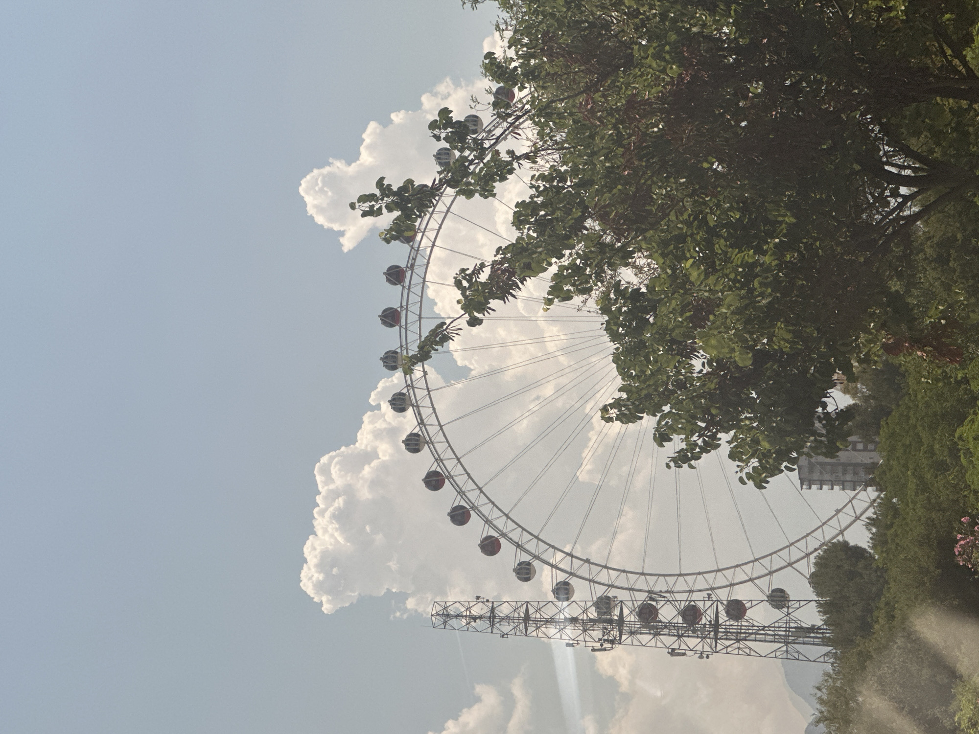 This image depicts a large Ferris wheel in Konyaaltı, Turkey. The Ferris wheel is the central focus, with its circular structure and numerous cabins visible. To the left of the Ferris wheel, a tall, lattice-like tower, possibly a transmission tower or part of the amusement park's infrastructure, stands prominently. The background features a bright blue sky with fluffy white clouds. In the foreground and midground, lush green trees partially obscure the Ferris wheel, adding a natural element to the scene. A building can be seen behind the Ferris wheel and trees, suggesting an urban or semi-urban setting. The overall impression is of an amusement park or recreational area.