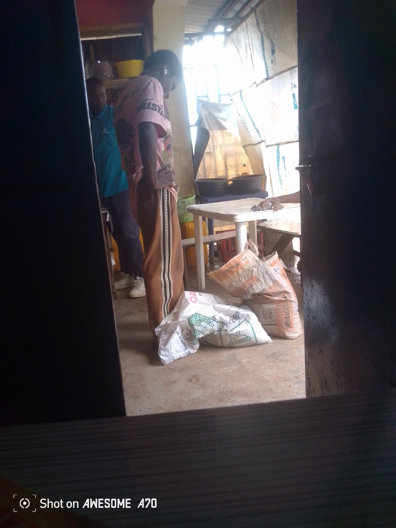 A person wearing a pink t-shirt and brown pants stands in a room. The person has their hands on their hips and is looking down. In the background, another person wearing a blue shirt and dark pants stands near a shelf. The room appears to be a storage area or market, with bags of what appear to be goods on the floor. There is also a table and chairs in the room. The overall lighting is dim, with natural light coming from a window or opening in the background.