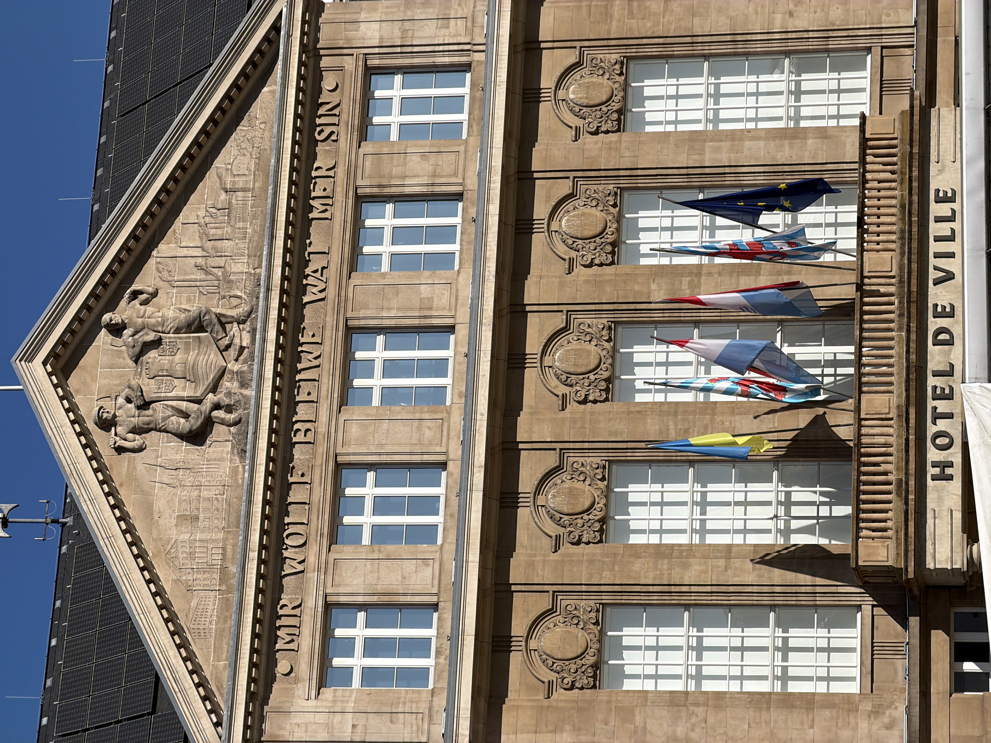 An upward-angled, sunlit view captures the facade of a historical stone building, identified as "HOTEL DE VILLE" (City Hall) by vertical text on a right-side pillar. The building, located in Esch-sur-Alzette, Luxembourg, is constructed from light brown/tan stone with numerous white-framed windows arranged in multiple rows.
On the left side of the facade, a large triangular pediment features an elaborate stone relief sculpture. This carving depicts figures, including a reclining male figure at the bottom, possibly representing a worker or miner, along with what appears to be a coat of arms or shield. To the left, another figure is seen holding a tool. Running vertically alongside this section is carved text: "OMR WOLLE BLEIWE WAT MER SIN C," a partial rendering of the national motto of Luxembourg, "Mir wëlle bleiwe wat mir sinn" (We want to remain what we are).
Between and around the windows, the building is adorned with ornate, carved stone decorative elements, some featuring intricate scrollwork. A striking modern art installation is suspended in front of the windows, consisting of several colorful, stylized aircraft or kite-like objects. These include designs resembling the European Union flag (dark blue with yellow stars) and others in red, white, and blue patterns, possibly evoking the Luxembourgish flag.
To the far left of the historic building, a modern structure with dark, gridded panels, likely solar panels, is partially visible against a clear, bright blue sky. The strong natural sunlight casts distinct shadows, indicating a sunny day.