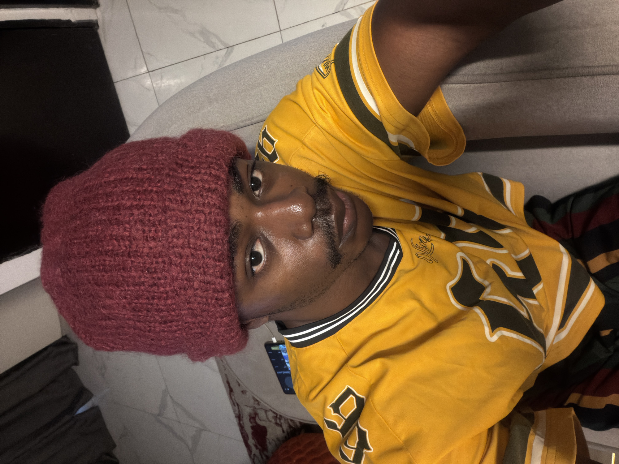 A person wearing a maroon knitted beanie and a yellow jersey with green and white stripes is captured in a close-up selfie. The background consists of a tiled floor, a gray couch, and curtains. A mobile phone is visible on the couch behind the person. The image appears to be taken indoors.