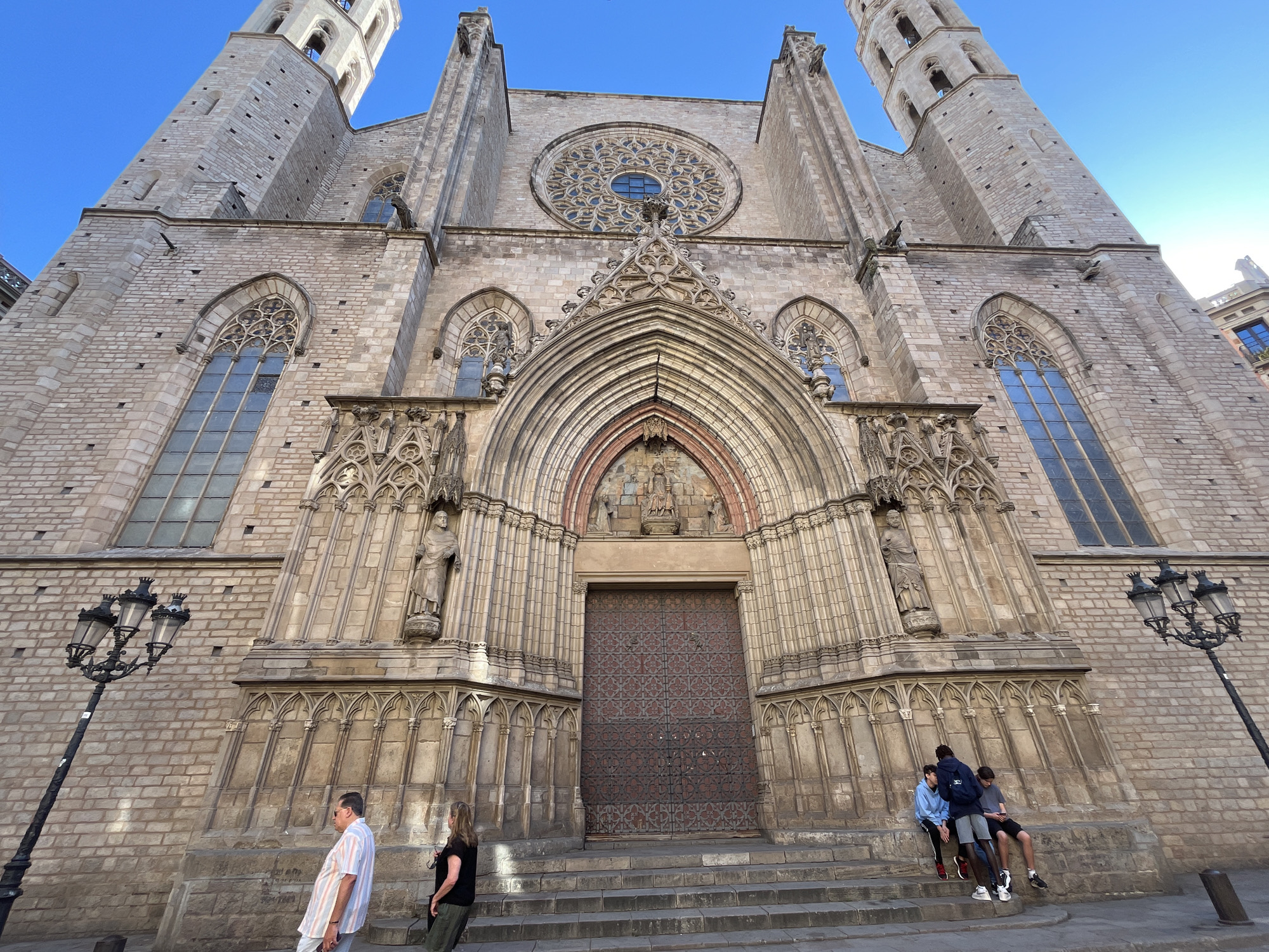 The image shows the facade of the Basilica de Santa Maria del Mar in Barcelona, Spain. The Gothic-style church features two tall towers, a large rose window above the main entrance, and ornate details around the arched portal.
In front of the church, several people are visible. On the left, a man in a striped shirt and a woman in dark clothing are walking past. On the right, three young people are sitting on the steps leading up to the main entrance. Two ornate street lamps flank the steps.
The image is taken from a low angle, emphasizing the height and grandeur of the church. The sky is clear and blue.