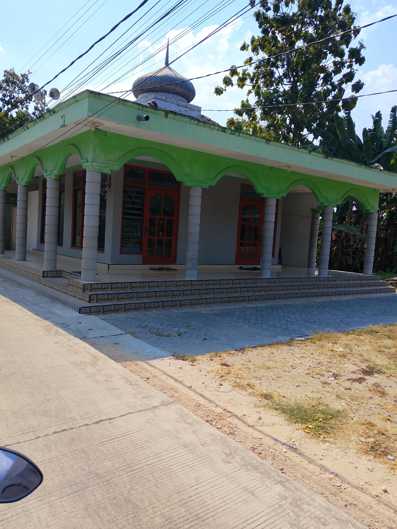 A mosque in Padangan, Indonesia, features a light green roof and a gray dome topped with a spire. The mosque has a covered porch supported by round pillars, with arched openings. Red doors and windows are visible. A tiled staircase leads up to the mosque. The ground in front of the mosque is paved with interlocking bricks, and a concrete path is to the left. Trees and vegetation are present in the background. The sky is blue with scattered clouds. There are no people visible in the scene.