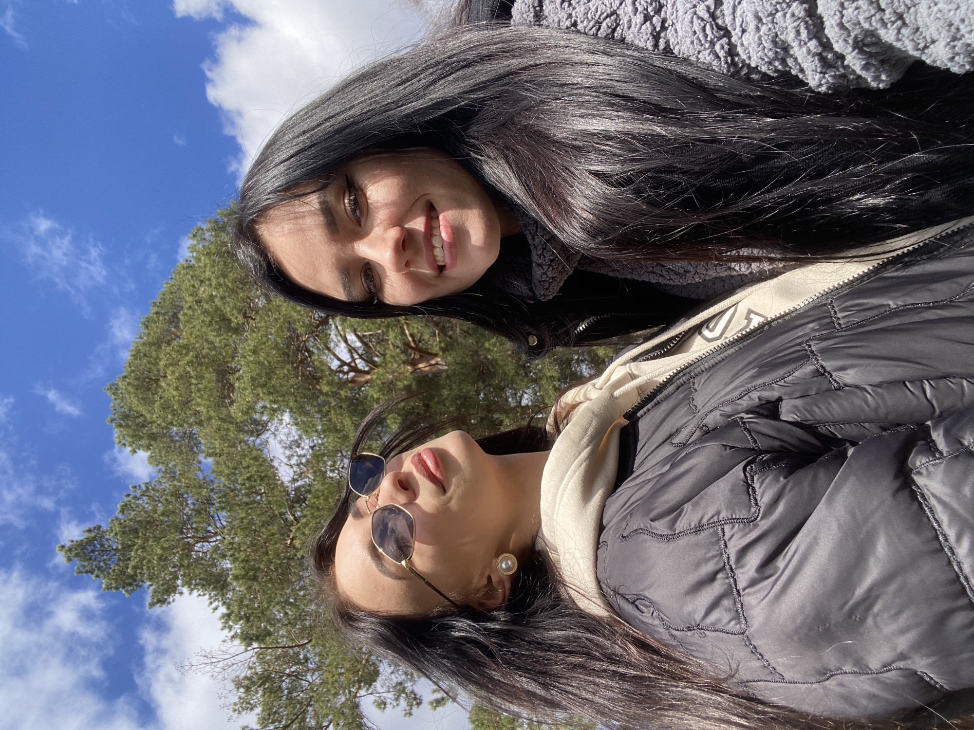 Two women are posing for a selfie outdoors. The woman on the top is smiling, wearing a gray sweater, with long dark hair. The woman below her is wearing sunglasses, a black puffy jacket with a beige hood, and an earring. They are in a park-like area in Chaikovsky, Russia. A large green tree and a blue sky with clouds are visible in the background.