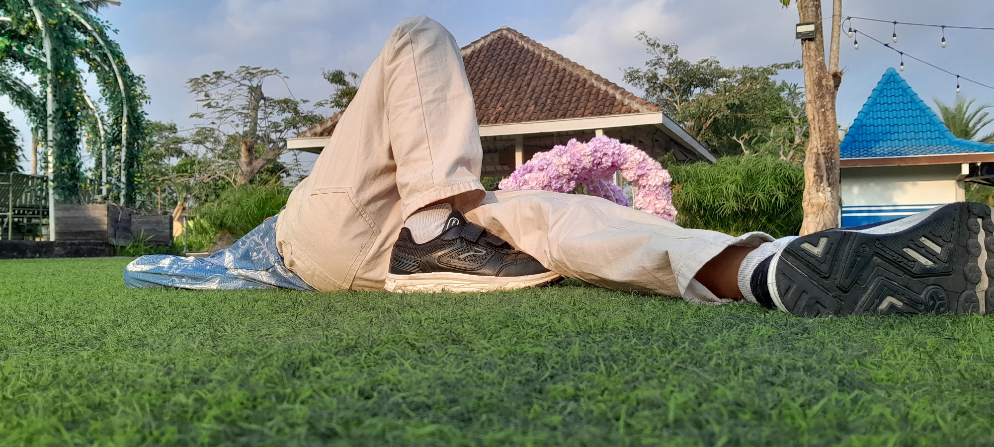 The image depicts a low-angle view of two pairs of legs resting on a vibrant green artificial turf lawn. The left person's legs are bent, with a blue and white patterned blanket draped over them. They are wearing light-colored pants and black sneakers with white soles and velcro straps. The right person's legs are also bent, with the feet pointing towards the right side of the frame. They are wearing light-colored pants and dark sneakers with white accents and white socks. In the background, a cluster of purple flowers forms an arch. Further back, a building with a red tiled roof and white walls is visible, along with a blue-roofed structure. Trees and foliage surround the area, and a clear sky with some clouds is above. String lights hang from a tree on the right side of the frame. This scene is set in Sleman, Indonesia, likely in an outdoor recreational or event space.