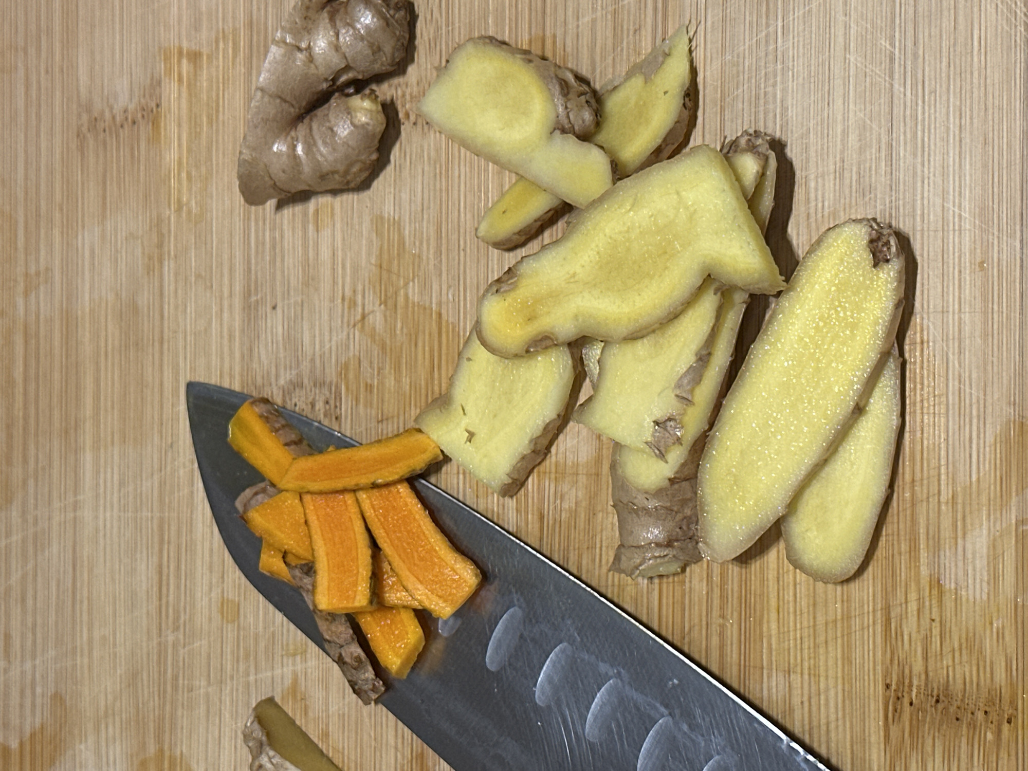 The scene depicts a wooden cutting board with sliced ginger and turmeric root. A silver knife lies diagonally across the board, with orange turmeric root slices on its blade. Scattered around are light yellow ginger slices and a small, unpeeled ginger root. The location, Milan, Italy, is not discernible from the image.