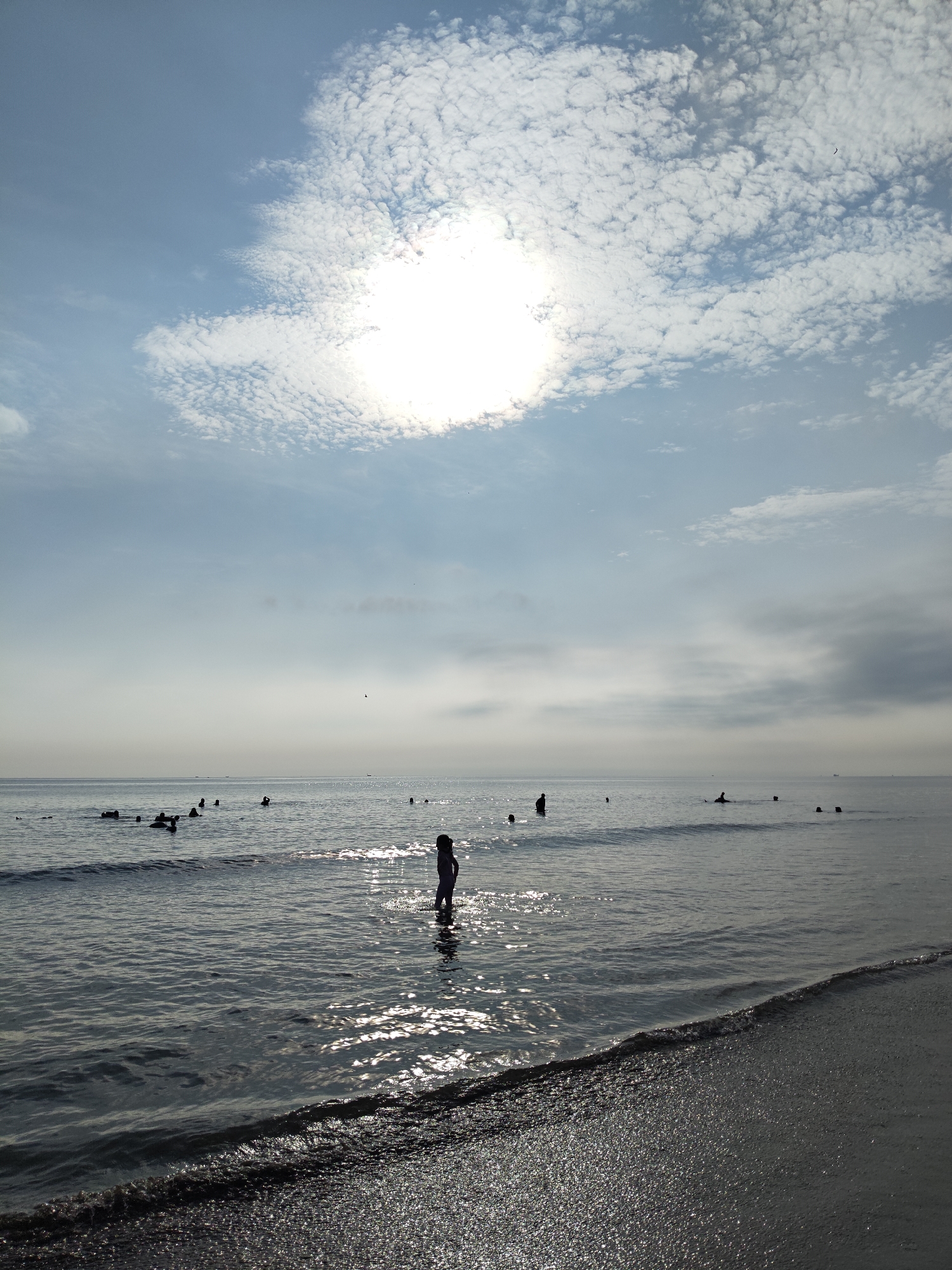 The image shows a beach scene in Lhokseumawe, Indonesia. The sky is partly cloudy with a bright sun. The sea is calm with gentle waves. Several people are in the water, some standing and others swimming. A person, seemingly a child, is standing in the shallows near the shore. The beach is visible in the foreground.