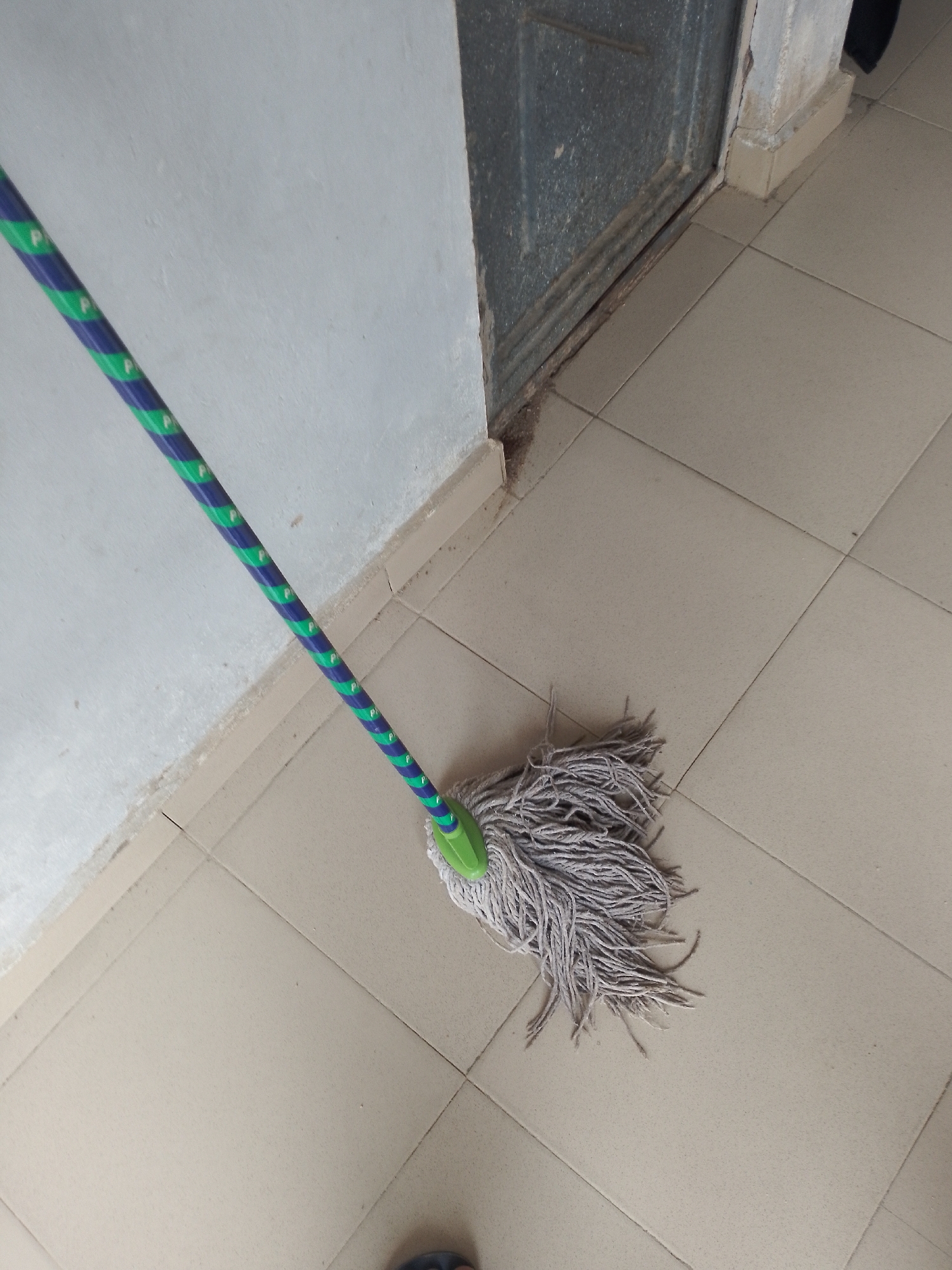 The image shows a mop with a blue and green striped handle resting on a tiled floor. The mop head is made of gray, frayed strings. To the left of the mop is a white wall, and to the right is a gray metal doorframe and a section of a gray, weathered door. The floor tiles are a light beige color and are arranged in a grid pattern. The scene appears to be indoors, possibly in a hallway or entryway, with no specific indicators of the city of Jauro Yinu, Nigeria. No people are clearly visible in the image, though a small portion of a dark-colored footwear is visible at the bottom edge.