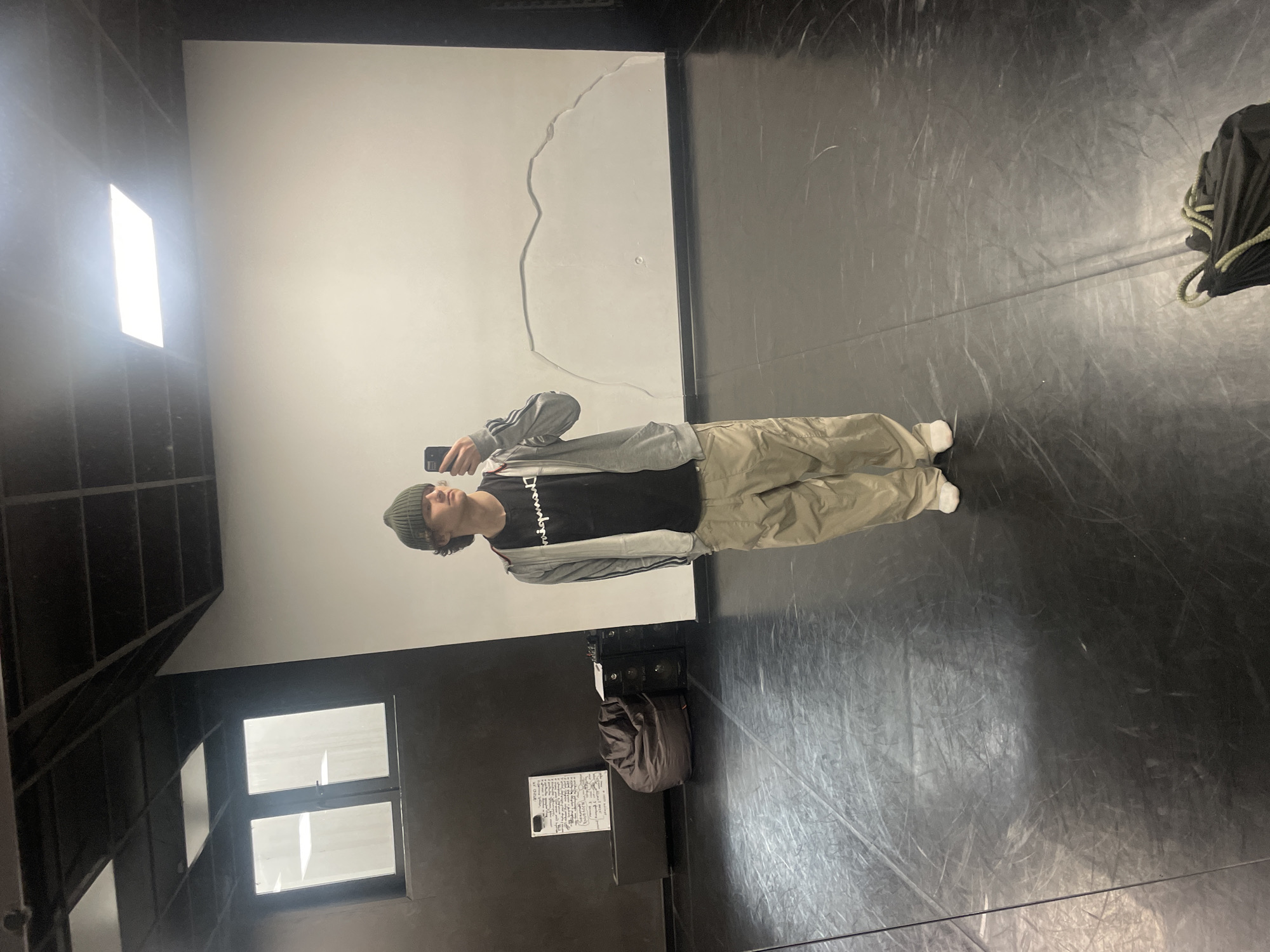 A young person takes a mirror selfie inside what appears to be a studio or functional indoor space, likely located in Krasnoyarsk, Russia. The individual, standing upright, is holding a dark smartphone to capture their reflection. They are dressed in casual attire, including a green beanie, a black t-shirt with "Champion" written on it, a grey hooded jacket with white stripes on the sleeves, baggy beige or khaki pants, and white sneakers.
The background features a large, plain white wall that serves as the main reflective surface for the selfie. A notable detail on this wall is a prominent, irregular crack or mark that extends vertically and then horizontally across its surface. The floor is dark, possibly a polished concrete or marley dance floor, showing numerous scuff marks and reflections.
To the person's left, a bright, rectangular light source or window is visible, alongside dark, grid-like structural elements. Below this, a smaller window with two panes can be seen. On the right side of the image, near the floor, a dark, soft bag (possibly a duffel or backpack) is present, accompanied by some coiled rope. Further to the left, on a dark counter or surface, a piece of paper with handwritten text is visible, though the text itself is illegible. The overall environment suggests a utilitarian or artistic space.
