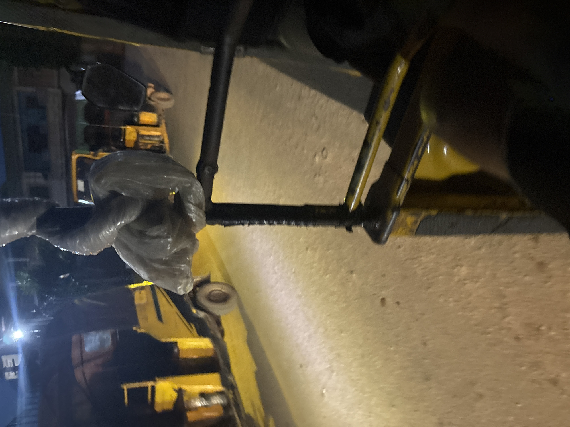 The image depicts a nighttime street scene in Onitsha, Nigeria. The foreground is dominated by the interior of a vehicle, likely a passenger auto-rickshaw, with parts of its yellow body visible. A plastic-wrapped object, possibly luggage or a bundle, is tied to a metal bar. The street outside is illuminated by artificial lights, casting long shadows. Several auto-rickshaws are parked or moving on the road, their yellow and green bodies contrasting with the dark surroundings. Buildings, dimly lit, line the street in the background. The overall impression is that of a bustling urban environment at night, with typical modes of transport.