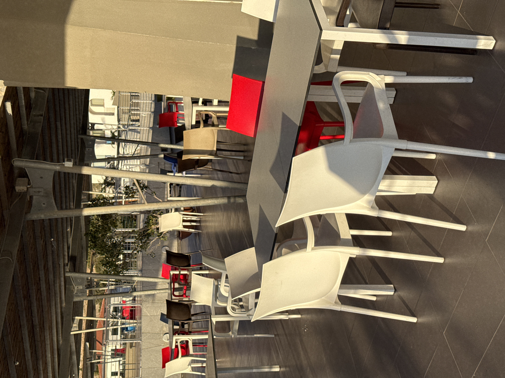 An outdoor seating area is depicted, likely a cafe or restaurant, under a shaded canopy with visible metal supports and a wooden slatted ceiling. The floor is paved with dark square tiles. Several tables and chairs are arranged throughout the space.
In the foreground, a large rectangular table with a gray surface is prominent. Two white chairs with arms are positioned at this table, one in the lower left and another in the lower right foreground, both facing inward. A red chair is visible on the far side of this table.
Further back, a variety of other tables and chairs are present. These include white, red, and dark-colored chairs, some with straight backs and others with more rounded designs. Some tables have red chair and some have black chairs. One table in the middle distance has a red chair and a dark brown chair. Another table has a red chair and a white chair.
In the background, beyond the immediate seating area, there's a glimpse of buildings with white walls and red-tiled roofs, suggesting a Mediterranean setting. A tree with green foliage is also visible. The sunlight casts long shadows across the tiled floor, indicating it is either early morning or late afternoon. The scene is calm and devoid of any people or activity.