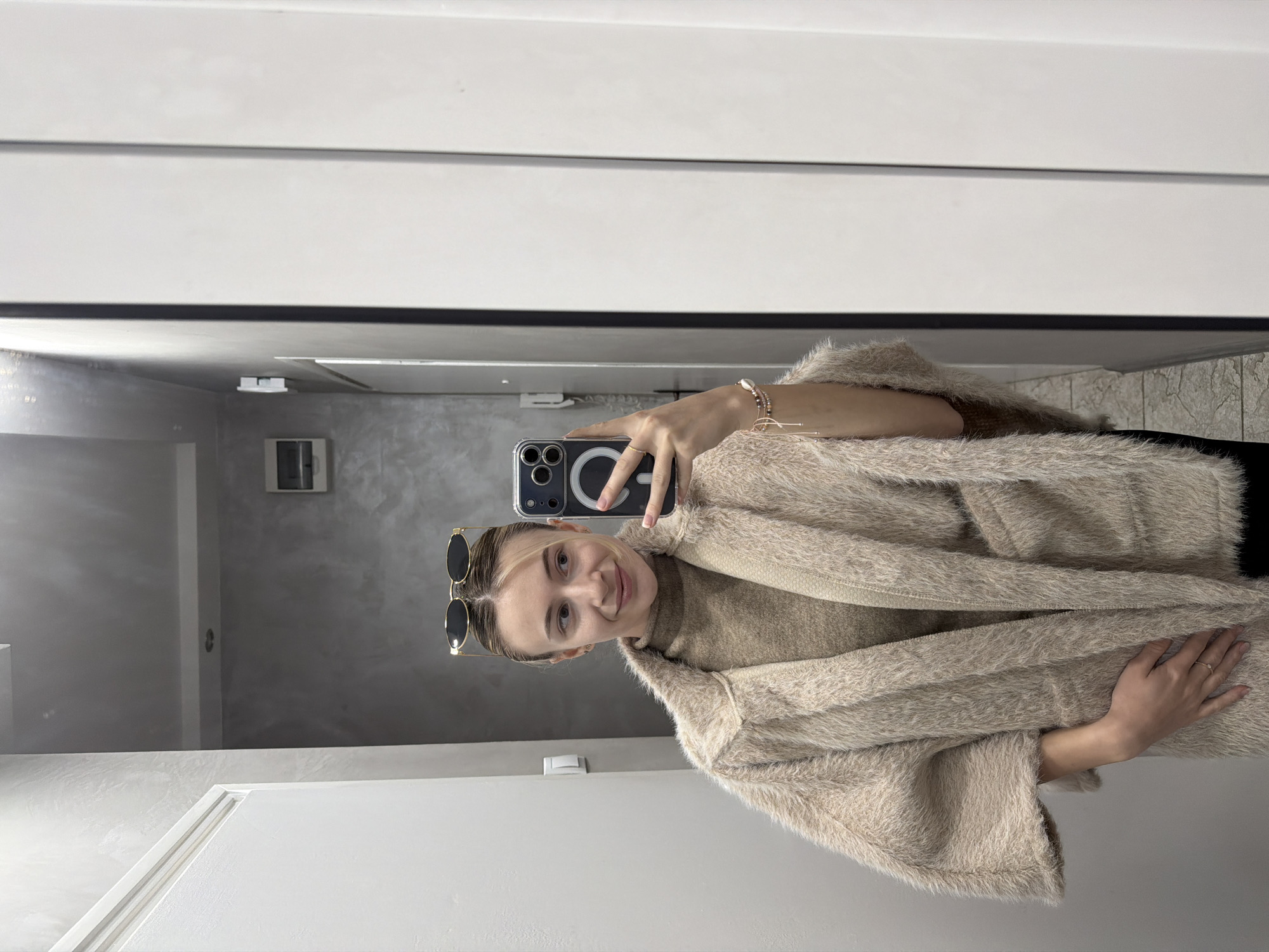 A young woman takes a selfie in front of a mirror. She has sunglasses perched on her head, is wearing a light brown turtleneck sweater and a fuzzy, light brown cardigan. She is holding her phone in her right hand, and her left hand is resting on the cardigan. The background appears to be a room with light gray walls and white trim. There is a doorway visible in the mirror's reflection. The location of this image in the city of Plovdiv, Bulgaria cannot be confirmed from the given image.