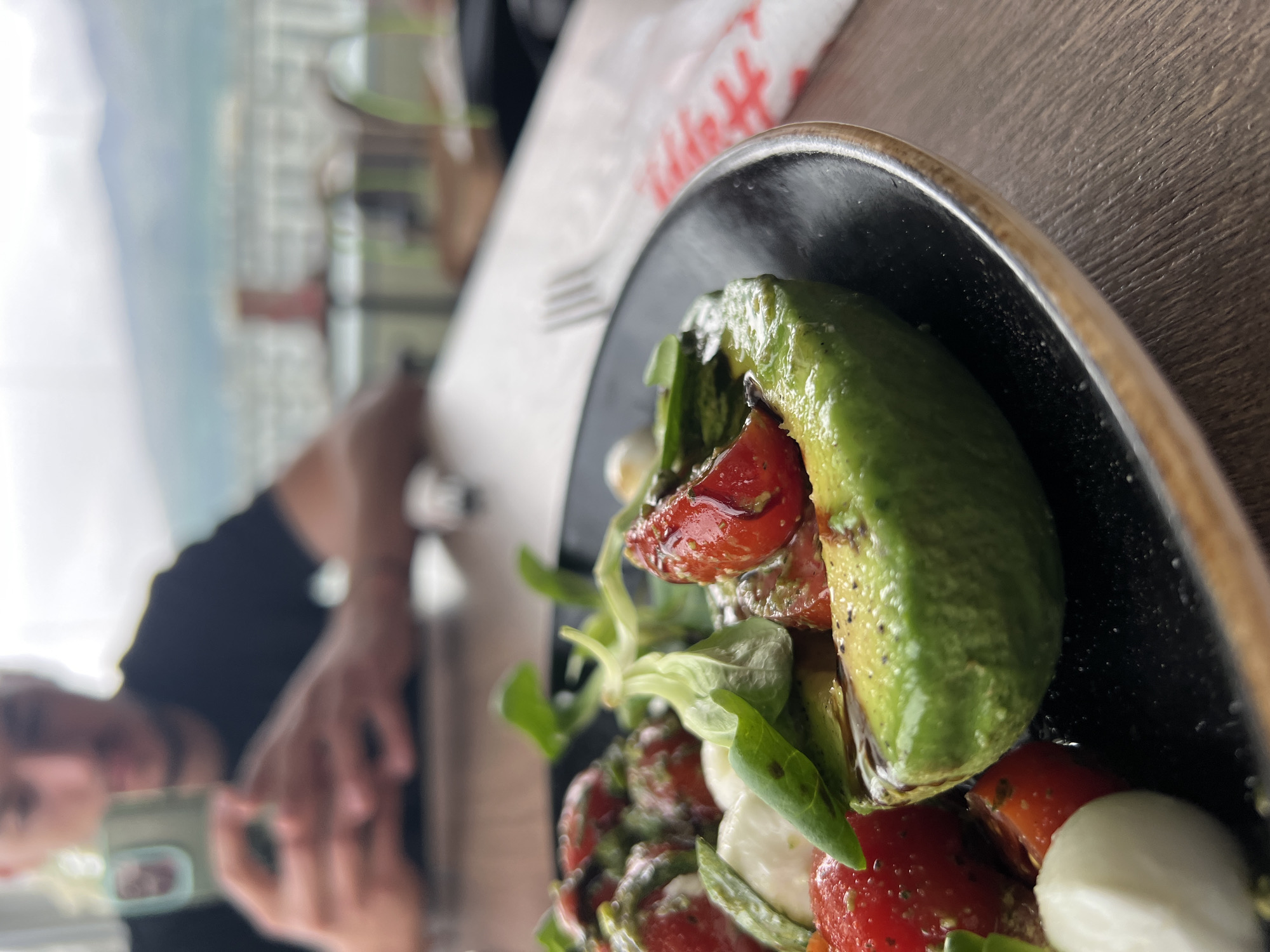 The image shows a close-up of a salad in a black bowl with a gold rim, consisting of avocado, tomatoes, mozzarella, and green leaves, possibly basil, seasoned with a sauce. A fork is resting on the table behind the bowl. In the blurred background, a person is holding a phone and may be taking the photo. The image appears to be taken inside a restaurant with a blurred cityscape visible in the background. Based on the architectural style visible in the background, the image could possibly be taken in Sofia, Bulgaria. The table surface is wooden. A white paper with red text is also partially visible on the table near the bowl.