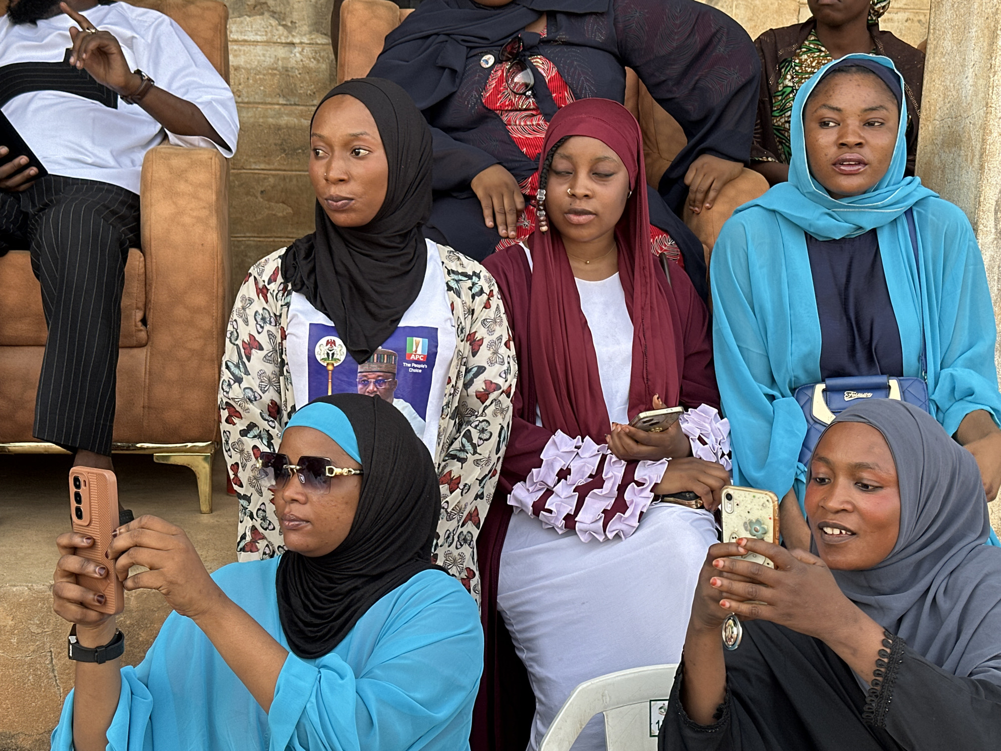 The image shows a group of six women and one man, possibly at an outdoor event in the city of Unknown, Unknown. The women are wearing hijabs and various colorful dresses and blouses. Two of the women in the foreground are holding and looking at their smartphones, possibly taking photos or videos. One woman has sunglasses. A third woman is looking at her phone, but not holding it up to record. In the background, more women are sitting. The man is wearing a white shirt and black pinstripe pants and is sitting on a brown cushioned seat. The setting appears to be a public space with tan colored concrete. The people are seated in chairs.