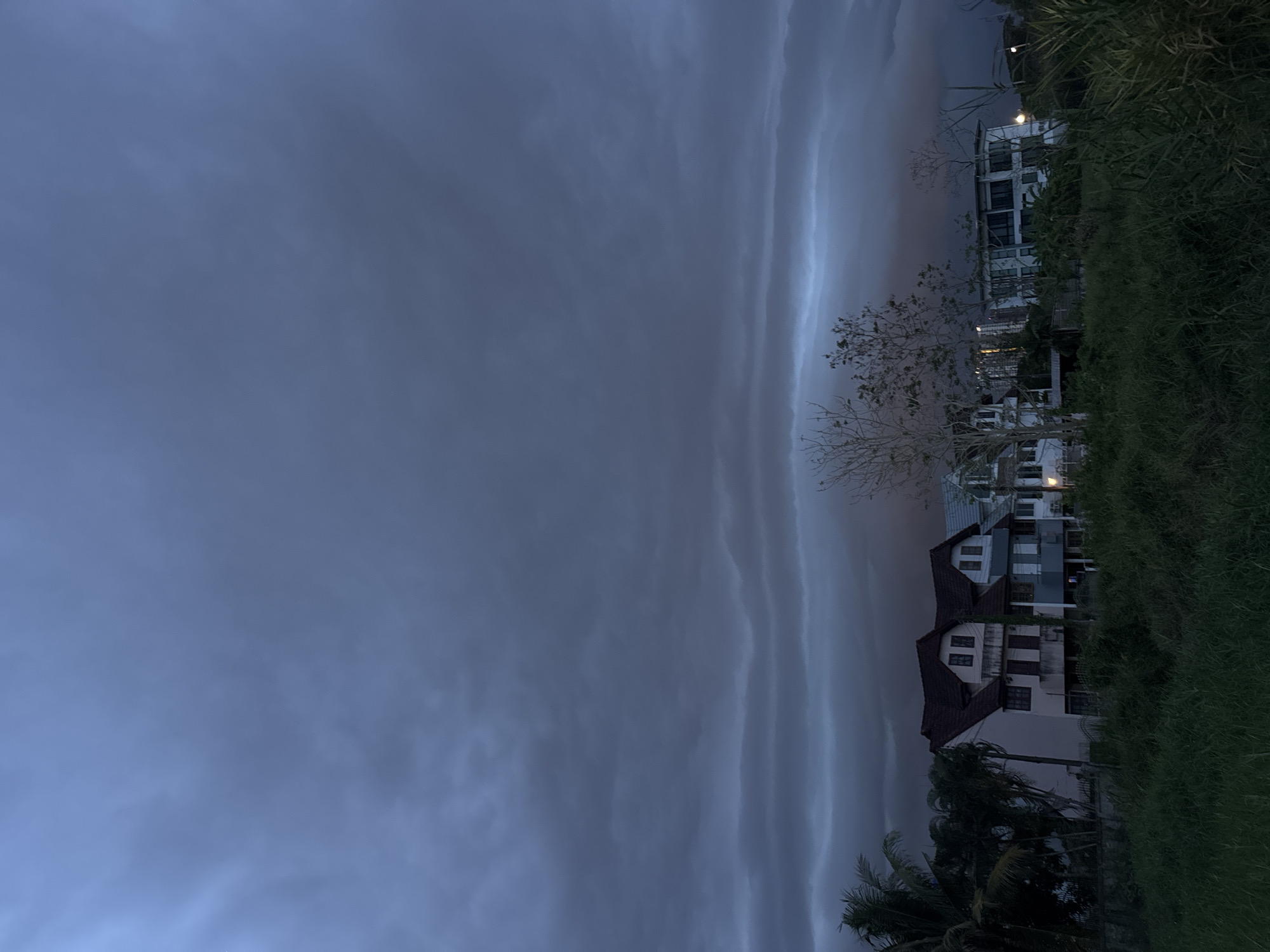 The image shows a view from บ้านกาญจน์กนก 23, Thailand, with a focus on the sky and nearby buildings. The sky is dominated by dark, ominous clouds with distinct striated patterns. Several multi-story buildings are visible on the right side of the frame, interspersed with trees and lush green vegetation. The scene is dimly lit, suggesting either twilight or overcast conditions.