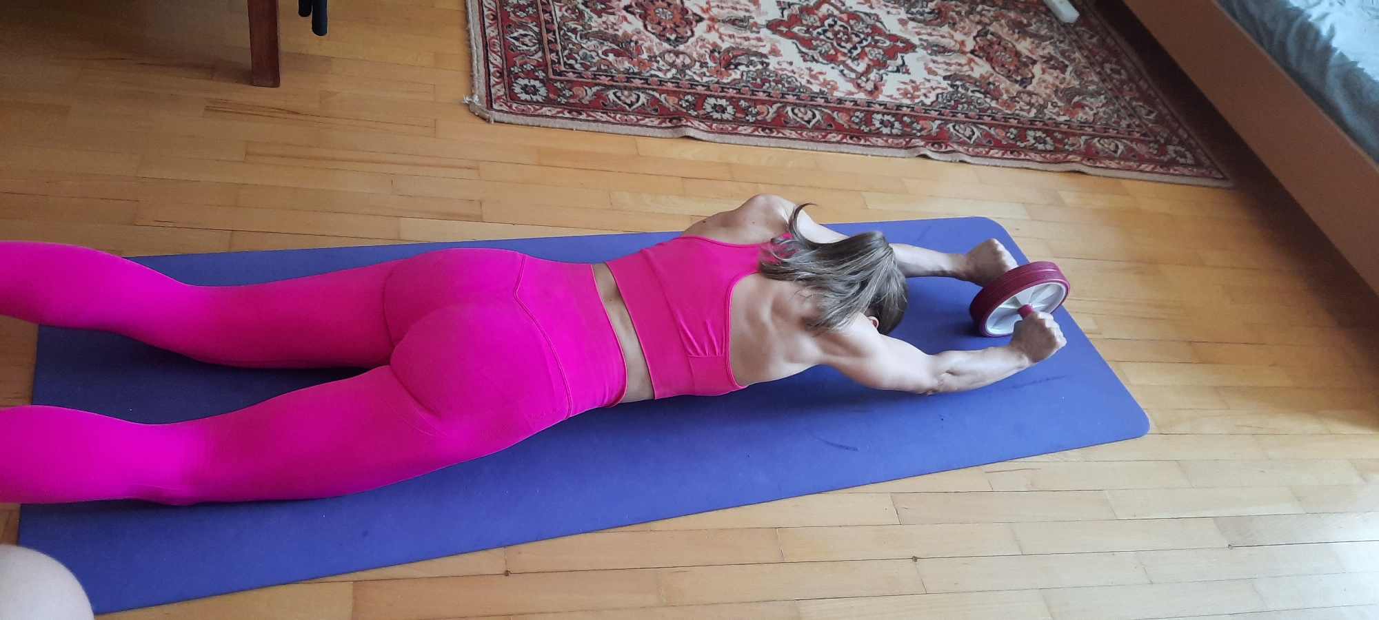 An overhead indoor shot captures a person, likely female, engaged in an exercise using an abdominal roller. The individual is lying face down on a blue exercise mat, fully extended, with both hands grasping a red and white ab roller. She is dressed in a bright pink sports bra and matching pink leggings. Her back and arms show visible muscle definition. Brown or dark blonde hair is partially visible, falling forward. The setting appears to be a home in Moscow, Russia, featuring a light wooden floor, a traditional patterned rug with red, cream, and blue designs, and glimpses of furniture including a wooden table leg and a bed frame. The scene is brightly lit, suggesting daytime. The person is performing a core strength exercise. No text is visible in the image.