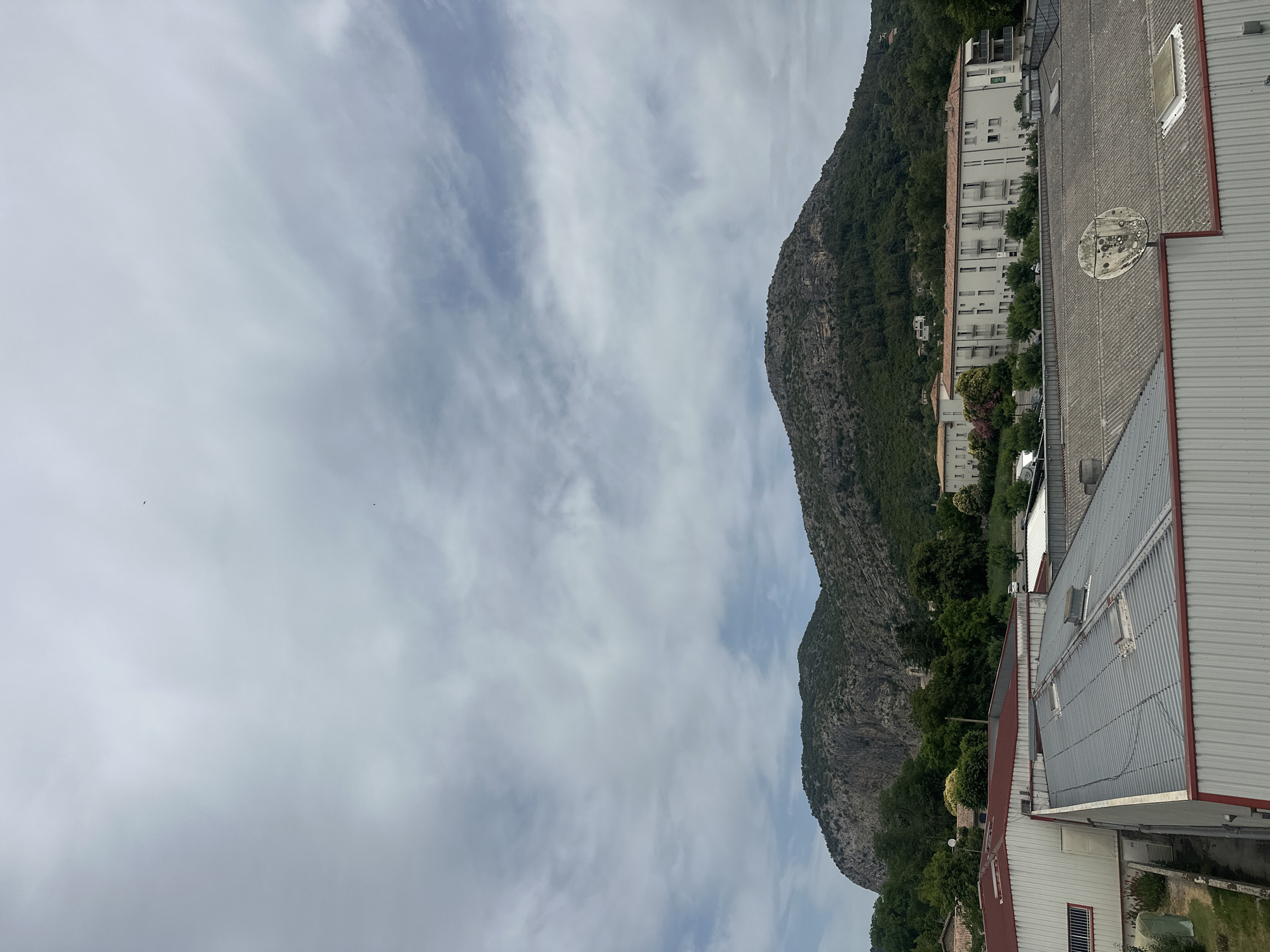 This media file shows a view from a rooftop in Anduze, France. The scene features a steep, rocky mountain covered in dense green vegetation on the left side of the image. The mountain slopes down towards a cluster of buildings situated at its base. Among these buildings, a white multi-story structure with numerous windows and balconies is prominent. In the foreground, a corrugated metal roof of an industrial building is visible, along with parts of other adjacent buildings. A satellite dish is mounted on the roof in the middle ground.
The sky is overcast with wispy clouds, suggesting a cloudy day. The lighting indicates it is daytime, likely late morning or early afternoon. The overall impression is of a somewhat industrial or commercial area nestled against a natural mountainous landscape. There is no visible text in the image. The image appears to be taken from a high vantage point, looking outwards and downwards towards the town and the mountain. The scene does not depict any people or specific activities, focusing instead on the architecture and the natural environment.