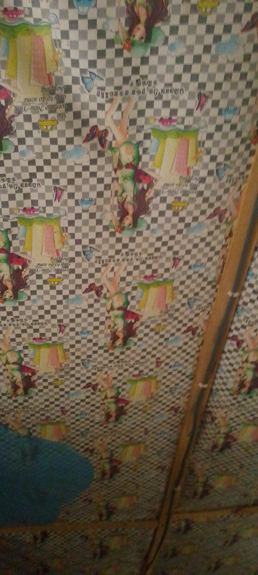 This is a close-up, oblique shot of wallpaper with a checkered pattern and numerous illustrations of cartoon-like figures, possibly anime-inspired, in various poses. The figures appear to be young women, some upside down, wearing colorful outfits and often depicted near stylized representations of clothing racks, clouds, or butterflies. There are also handwritten text elements scattered throughout, such as "She went to New York only to do some shopping." and "But hey, when do you really". The wallpaper is attached to a wall or ceiling, and a wooden beam or support structure runs vertically on the right side of the image. The lighting is dim, and there is a blurred blue object on the lower left, potentially a piece of furniture or fabric. The image does not provide any specific information about the city of Jalingo, Nigeria; it appears to be a generic interior scene.