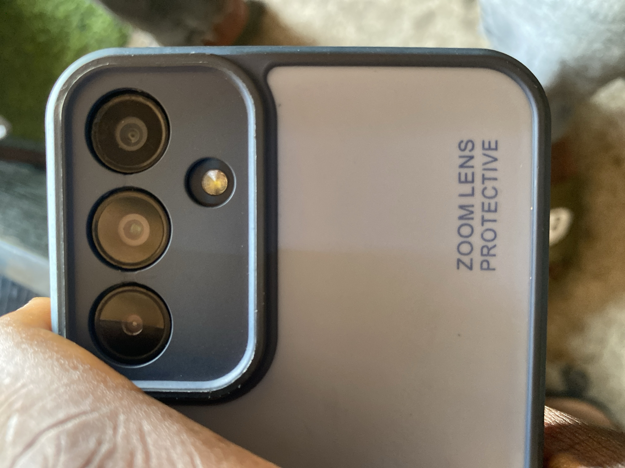 The image shows a close-up view of the camera module of a smartphone, encased in a protective cover. The camera module features three circular lenses arranged in a row at the top, with a smaller circular flash or sensor positioned below them. The cover is a dark grey color with a subtle sheen. Below the camera module, the words "ZOOM LENS PROTECTIVE" are printed in blue capital letters on the light grey back of the phone case. The background is blurred, but appears to be an outdoor setting with hints of green foliage and a rough, earthy surface, suggesting a natural or possibly urban environment. There is a person's finger visible at the top left of the image, holding the phone. The location in Howayi, Nigeria is not discernable from the image itself.
