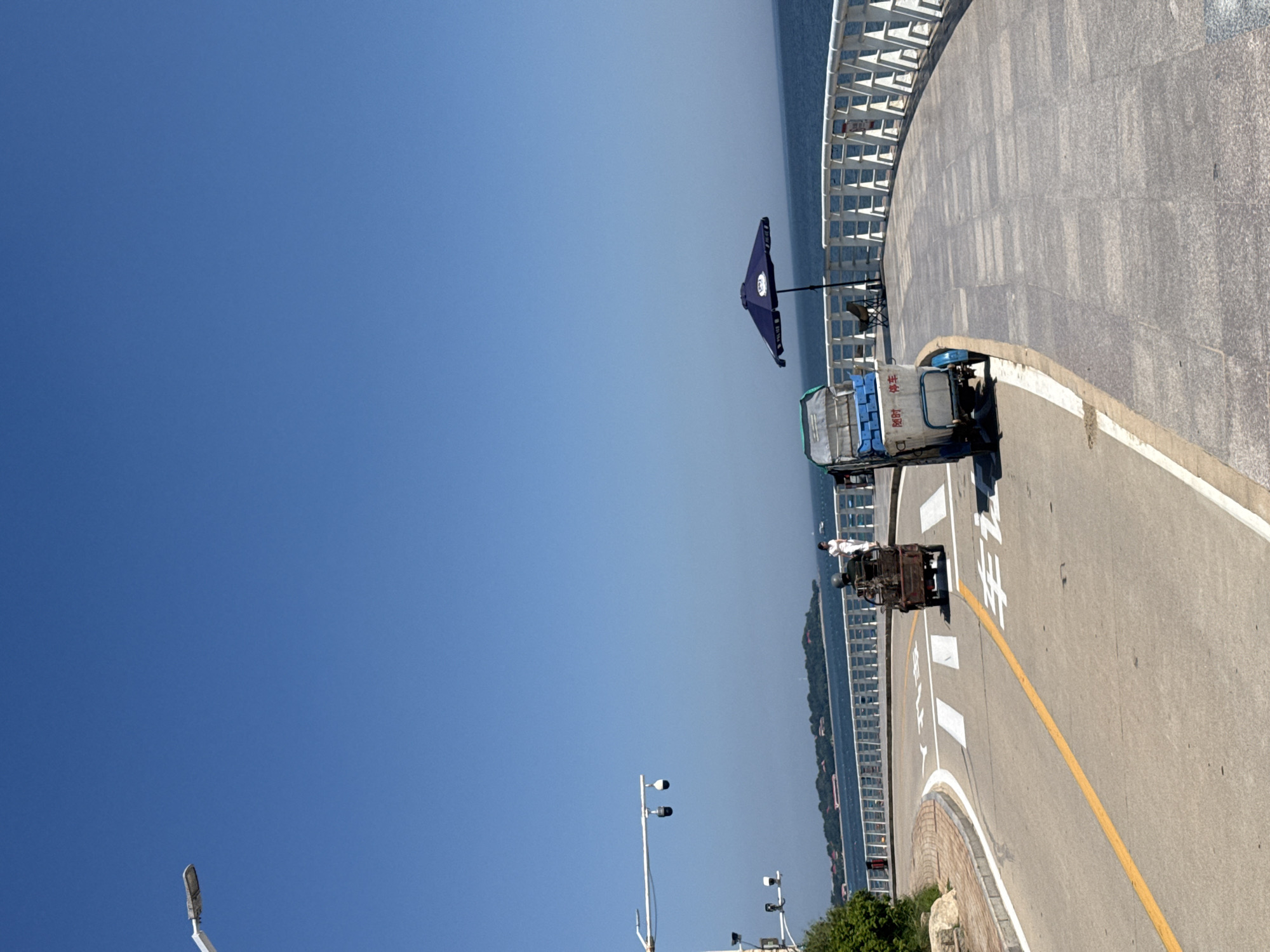 This image captures a sunny day along a coastal road in Qinhuangdao, China. The road curves to the right, with a yellow dividing line and white pedestrian crossing markings. On the left side of the road, a white streetlight with two cameras is visible against a clear blue sky. In the distance, across the sea, a tree-covered island with buildings can be seen.
To the right of the road, a white railing lines a promenade. A dark blue umbrella shades a chair, and a small three-wheeled vehicle with a covered cargo area, potentially used for deliveries or services, is parked. Another similar vehicle, carrying what appears to be equipment, is moving across the pedestrian crossing on the road. A woman in white is walking on the promenade. The sea stretches to the horizon, calm under the bright sun. The architecture suggests a coastal city environment, possibly a park or scenic route.