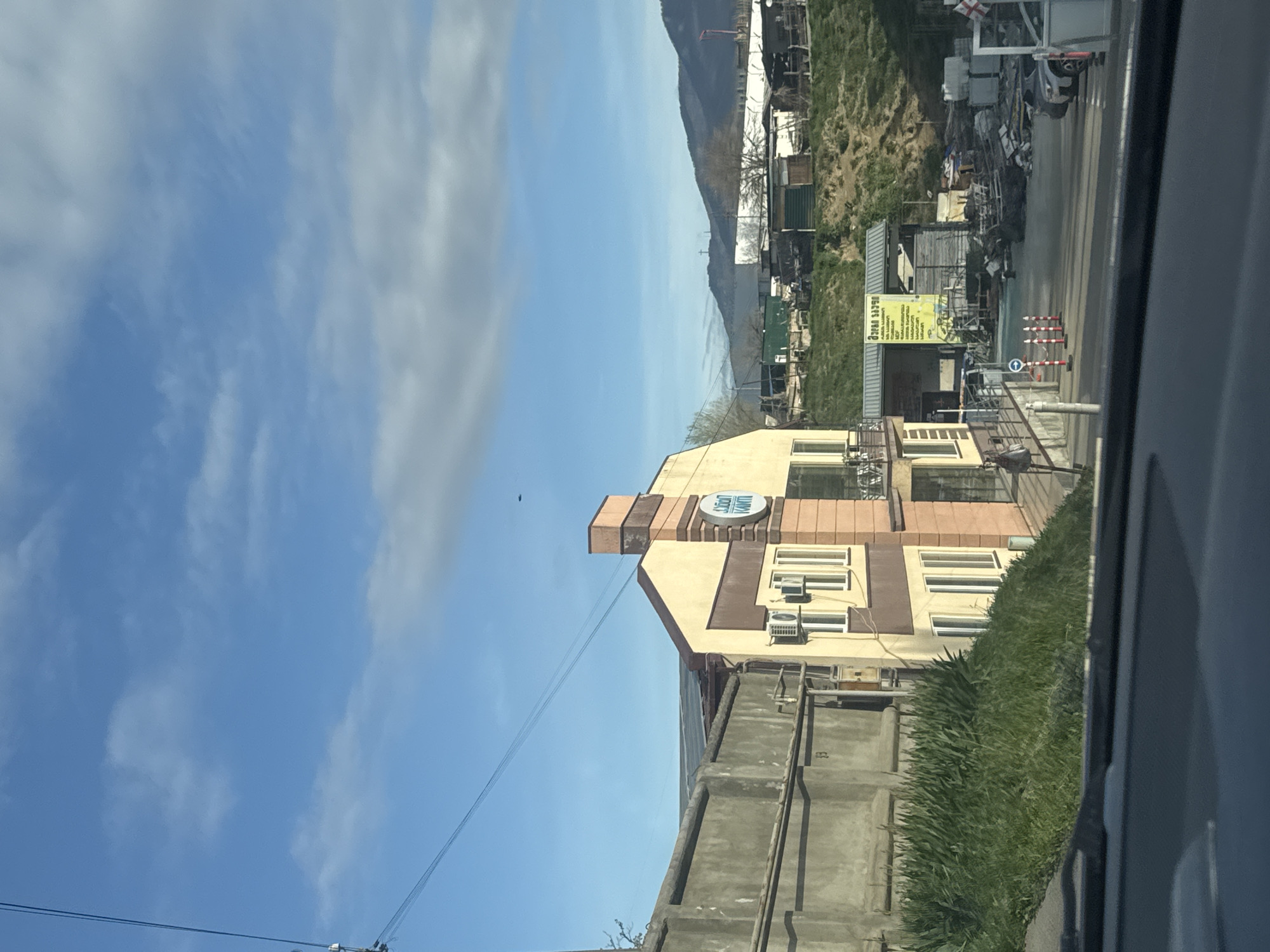 The image shows a view from inside a car, possibly moving, in Tbilisi, Georgia. The sky is blue with scattered clouds. A multi-story building with a beige and brown facade is prominent. The building has multiple windows and air conditioning units. A sign with text in Georgian is affixed to the building's upper portion. In the background are hills or low mountains and various industrial or commercial structures, including what appears to be a scrap yard or auto parts facility. There is a concrete wall with grass along its base in the foreground. Power lines stretch across the sky.