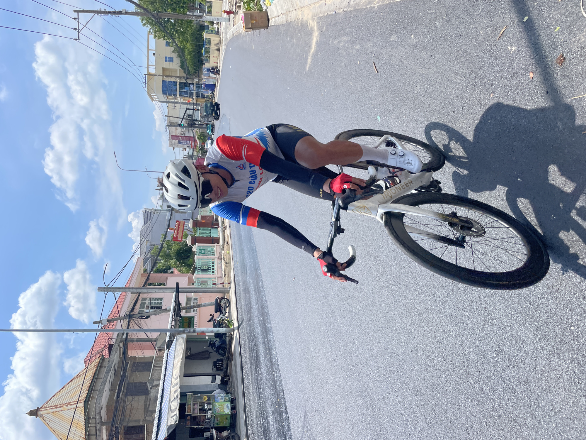 A male cyclist wearing a white helmet, sunglasses, and a cycling jersey with red and blue sleeves is riding a white road bike on a paved road in a city. He is also wearing black arm warmers, black cycling shorts, and white cycling shoes. The cyclist is smiling. The road appears to be recently paved, with some loose gravel. In the background, there are buildings, power lines, and trees. The sky is blue with white clouds. The scene is likely in the city of Phan Thiết, Vietnam, as indicated by the Vietnamese text on the cyclist's jersey.