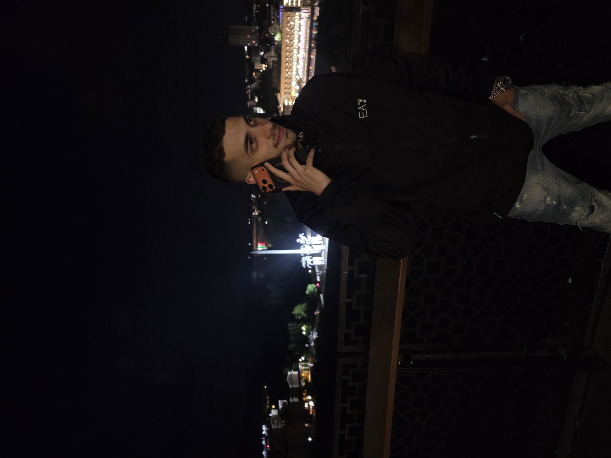A man is standing at night on a balcony in Veliko Tarnovo, Bulgaria, talking on his phone. He is wearing a dark jacket with the EA7 logo, distressed jeans, and a wristwatch. The illuminated cityscape is visible in the background. The railing of the balcony is in the foreground.