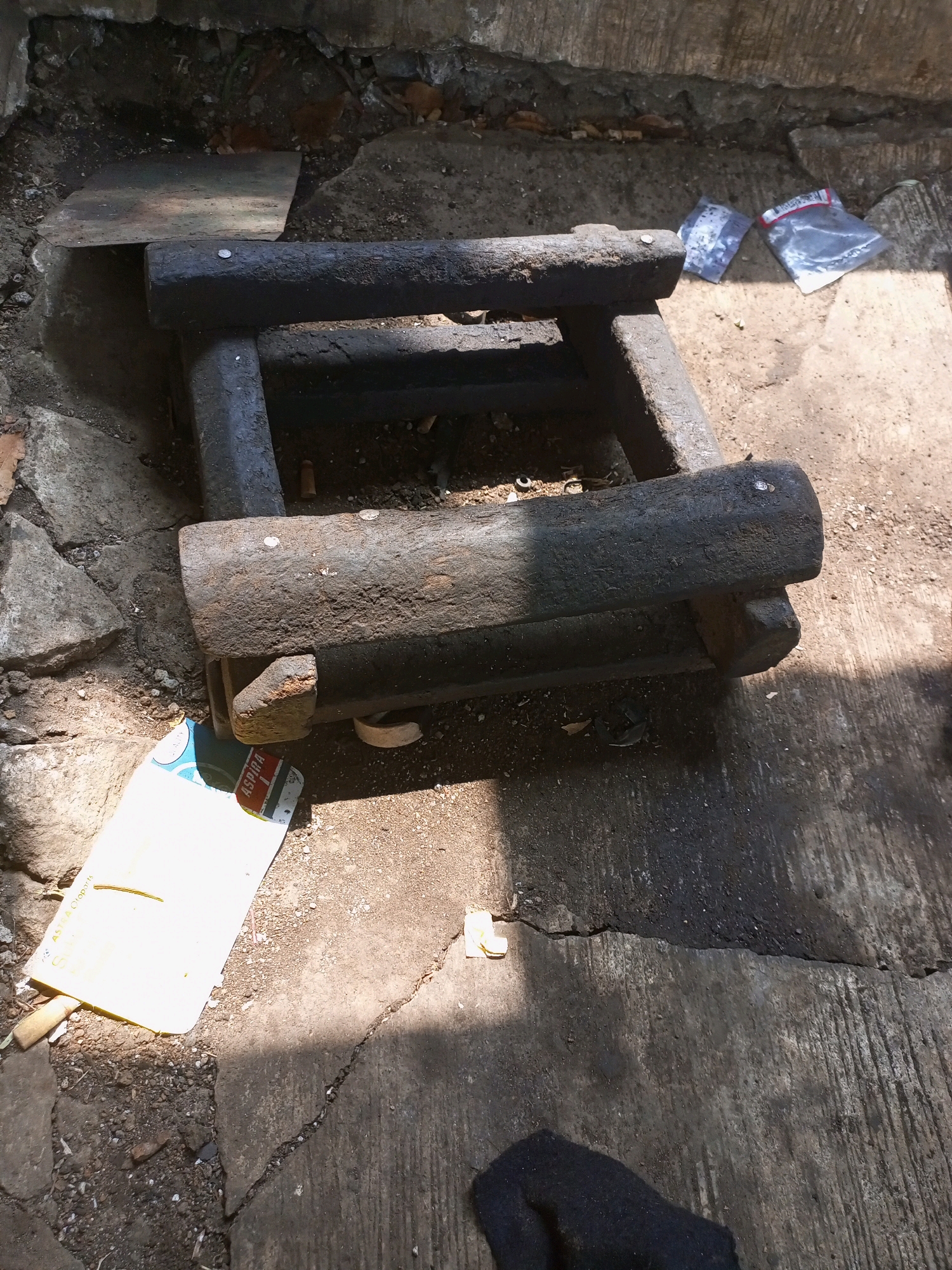 In Jatisrono, Indonesia, an outdoor scene depicts a dark, weathered wooden structure, possibly a primitive stool or bench, resting on cracked concrete and gravel. The object is composed of thick, rough-hewn logs, showing signs of age and use, with metal fasteners visible. To the left of the structure, a partially visible white and blue cardboard package with red lettering is placed on the ground. In the upper right quadrant of the image, two small, transparent plastic bags containing what appears to be small items are situated on the ground. The environment is illuminated by sunlight, casting shadows across the concrete surface, suggesting a daytime setting.