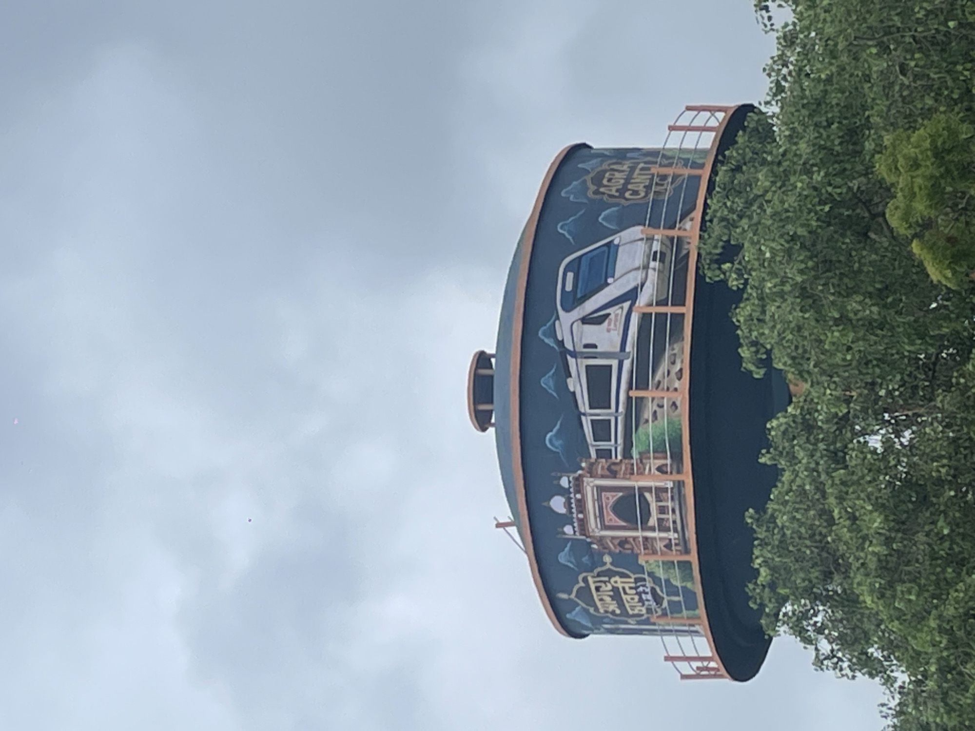 A circular water tower in Agra, India, is painted with a mural depicting a modern train and an Indian architectural monument, possibly the Taj Mahal, with text in Hindi and English. The text in Hindi reads "आगरा छावनी" which translates to "Agra Cantt" or "Agra Cantonment". The text in English reads "AGRA CANTT". The mural is bordered by a railing and surrounded by green trees, with a cloudy sky in the background. The overall scene is a public artwork integrated into the urban infrastructure of Agra.