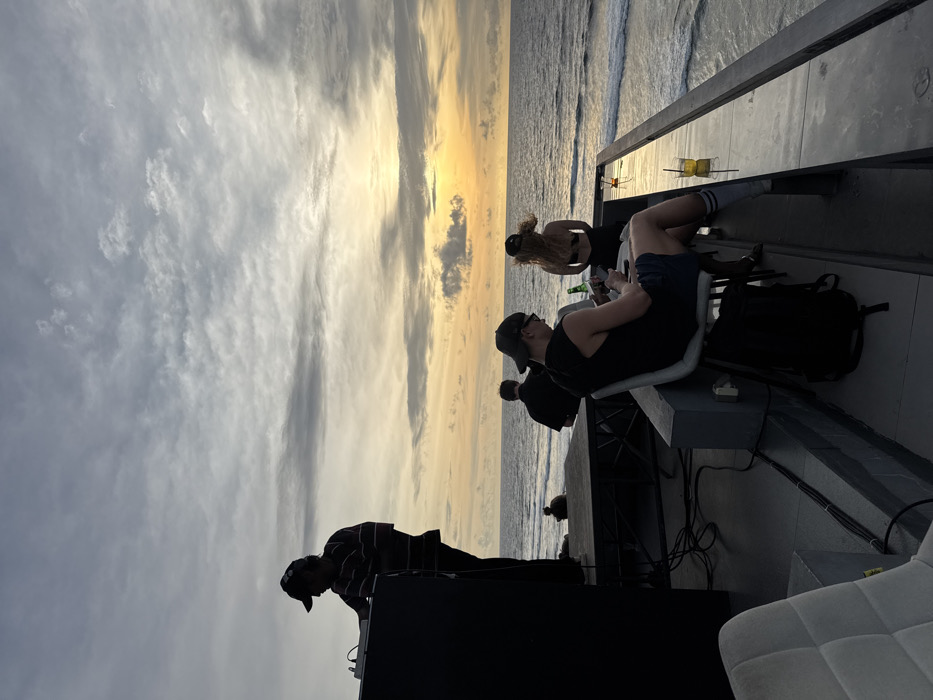 The image depicts an outdoor seaside venue in Uluwatu, Indonesia, at sunset. A male DJ, silhouetted against the sky, stands on a dark raised platform on the left, operating audio equipment with headphones on and wearing a cap and striped shirt. Cables are visible on the platform floor. To the right of the platform, four individuals are visible near a dark, reflective ledge overlooking the ocean. In the immediate foreground, a light-colored upholstered chair is partially visible.
A male in a dark tank top, dark shorts, sunglasses, and a backward baseball cap is seated on a light-colored chair in the midground, looking down at a smartphone. A dark backpack rests on the ground beside him. A tall glass containing a yellow liquid and a straw is on the ledge to his right. Behind him and slightly to the left, a female wearing a dark baseball cap and a dark bikini top stands facing the ocean, holding a green bottle. Another male is seated further left, facing away from the camera, observing the ocean. A fourth, more distant individual is seated or kneeling on the same elevated platform as the DJ, also facing the ocean. The ocean features gentle waves. The sky displays a clear gradient from an orange-yellow sunset glow at the horizon to a cloudy, muted grey-blue overhead.