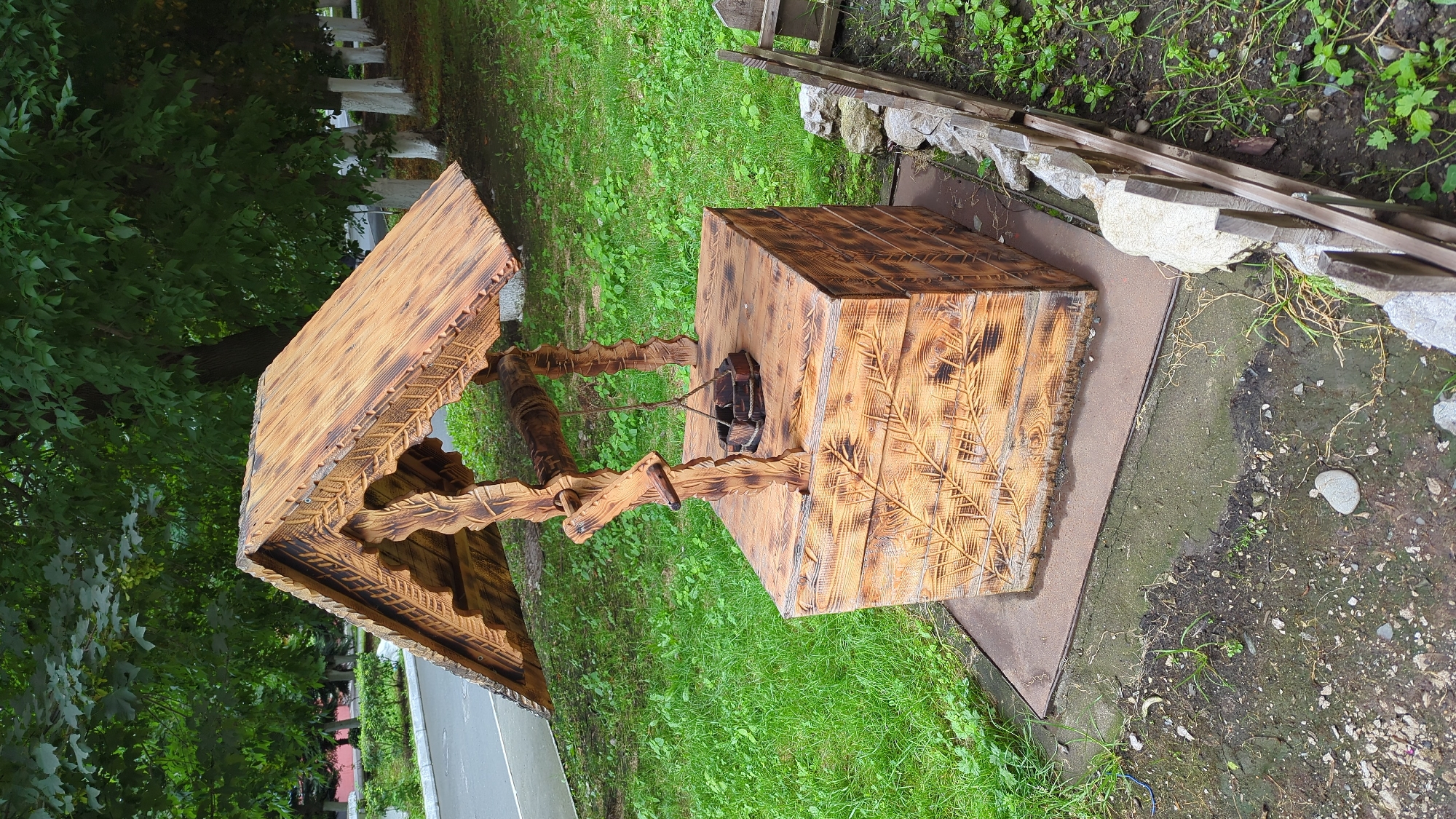 A decorative wooden well with a pitched roof sits on a square metal plate on a patch of grass. The well's base is a cube with decorative carvings, and the roof is supported by two carved wooden posts. A bucket hangs from a rope attached to a central beam. The well is positioned next to a simple wooden fence and surrounded by green grass. In the background, a row of trees with white-painted trunks lines a paved area. The scene appears to be in an outdoor, public space.