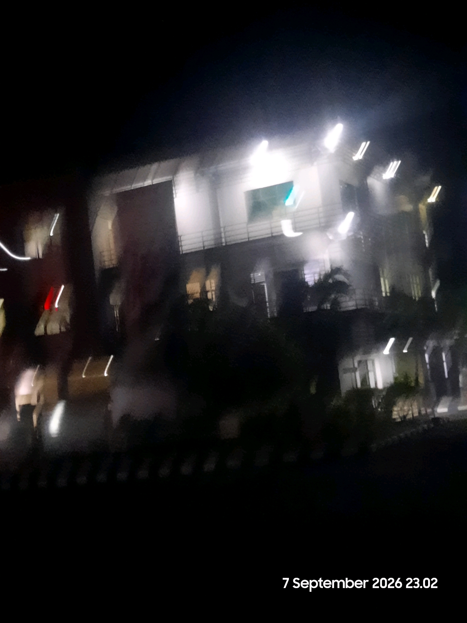 The image is a blurry nighttime photograph taken from a moving vehicle, likely a car. It shows a multi-story building with illuminated windows and balconies. The motion blur creates streaks of light from streetlights and building lights. In the foreground, there are some blurry figures or objects that are difficult to discern due to the low light and motion. The lower right corner of the image contains text that reads "7 September 2026 23.02", indicating the date and time the photograph was taken. There are no discernible details about the specific location within Gending, Indonesia, other than it appears to be an urban or semi-urban area with a building and outdoor lighting.