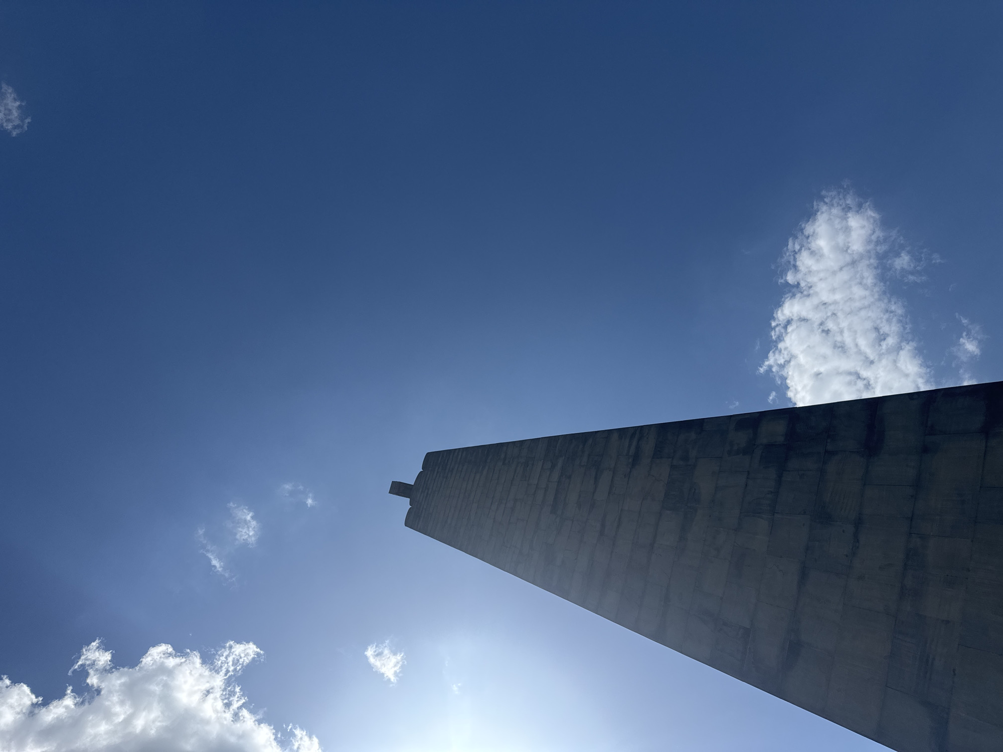 The image shows a low-angle view of the Mother Armenia statue in Yerevan, Armenia, under a clear blue sky with scattered clouds. The monument, constructed of grey stone blocks, is the dominant element in the frame, towering towards the upper right corner. The sun appears to be directly behind the base of the monument, casting a bright light around the lower portion. The sky is a gradient of blue, lighter near the horizon and deepening towards the top of the frame. Small, puffy white clouds are scattered across the sky, adding depth and texture to the scene.