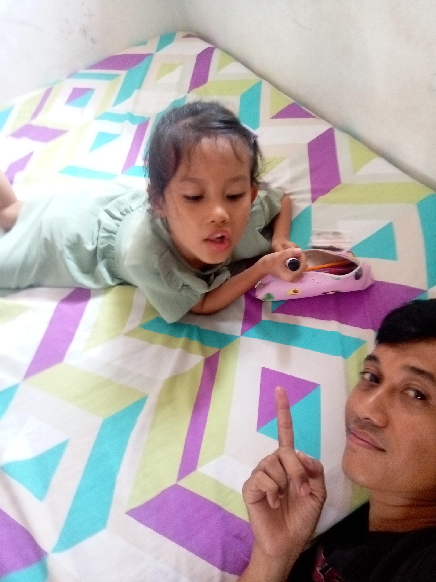 This media file shows two people, a young girl and a man, lying on a bed with a patterned bedsheet. The girl, on the left, is wearing a light green dress and is lying on her stomach. She has dark hair tied back and is looking towards the camera with her mouth slightly open. Next to her is a pink pencil case filled with colored pencils and a marker. The man is on the right, in the foreground, and is looking at the camera with a slight smile, holding up his right index finger. Both individuals appear to be in a relaxed and informal setting, likely a home. The bedsheet has a geometric pattern of overlapping rectangles and squares in shades of purple, light green, and turquoise on a white background. The lighting suggests it could be daytime, possibly indoors with natural light. The presence of a bed and the relaxed poses indicate a casual moment, perhaps a selfie taken by the man and girl. There is no visible text in the image.