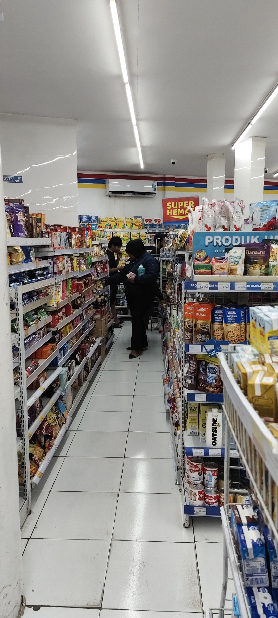 The image shows the interior of a convenience store, likely a minimarket in Malang, Indonesia. Two individuals are visible browsing the aisles. One person, wearing a dark hijab and black clothing, is standing with their back to the camera, holding a package and a bottle of hand sanitizer. Another person, wearing a black t-shirt and dark pants, is standing to the left of the first person, facing away from the camera and appearing to be eating. The store is well-lit with fluorescent lighting and features rows of shelves stocked with various products, including packaged foods, drinks, and household items. A "SUPER HEMAT" (Super Savings) sign is prominently displayed on the wall in the background. The floor is tiled in white. The overall scene depicts a typical retail environment.