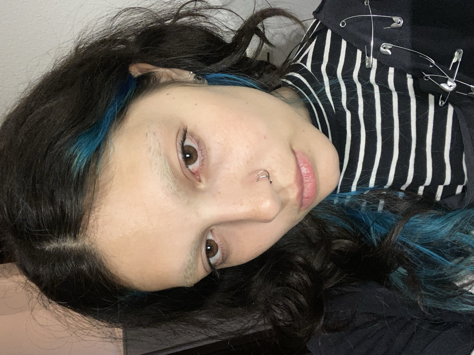 A young person lies on their left side, face directed towards the camera. The person has dark brown eyes, a nose ring, and dark hair with streaks of bright blue. They are wearing a black and white striped shirt adorned with safety pins. The background is a plain white wall and part of a dark object, potentially furniture. The location is not discernible from the image.