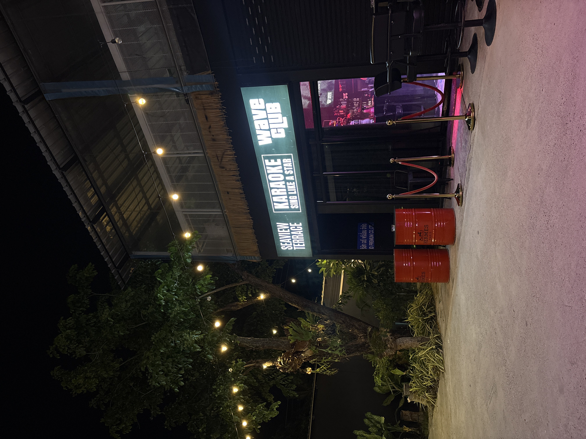 The image shows the entrance to the "Wave Club" karaoke bar in Baan Bang Rak, Thailand. The bar sign reads "SEAVIEW TERRACE KARAOKE, WAVE CLUB, SING LIKE A STAR." The entrance is marked by gold stanchions with red ropes. Two orange barrels, labeled "HERMES PARIS," are located near the entrance. A tree with string lights is in the foreground. A sign in Thai and English, "JD PREMIUM CO., LTD," is also visible.