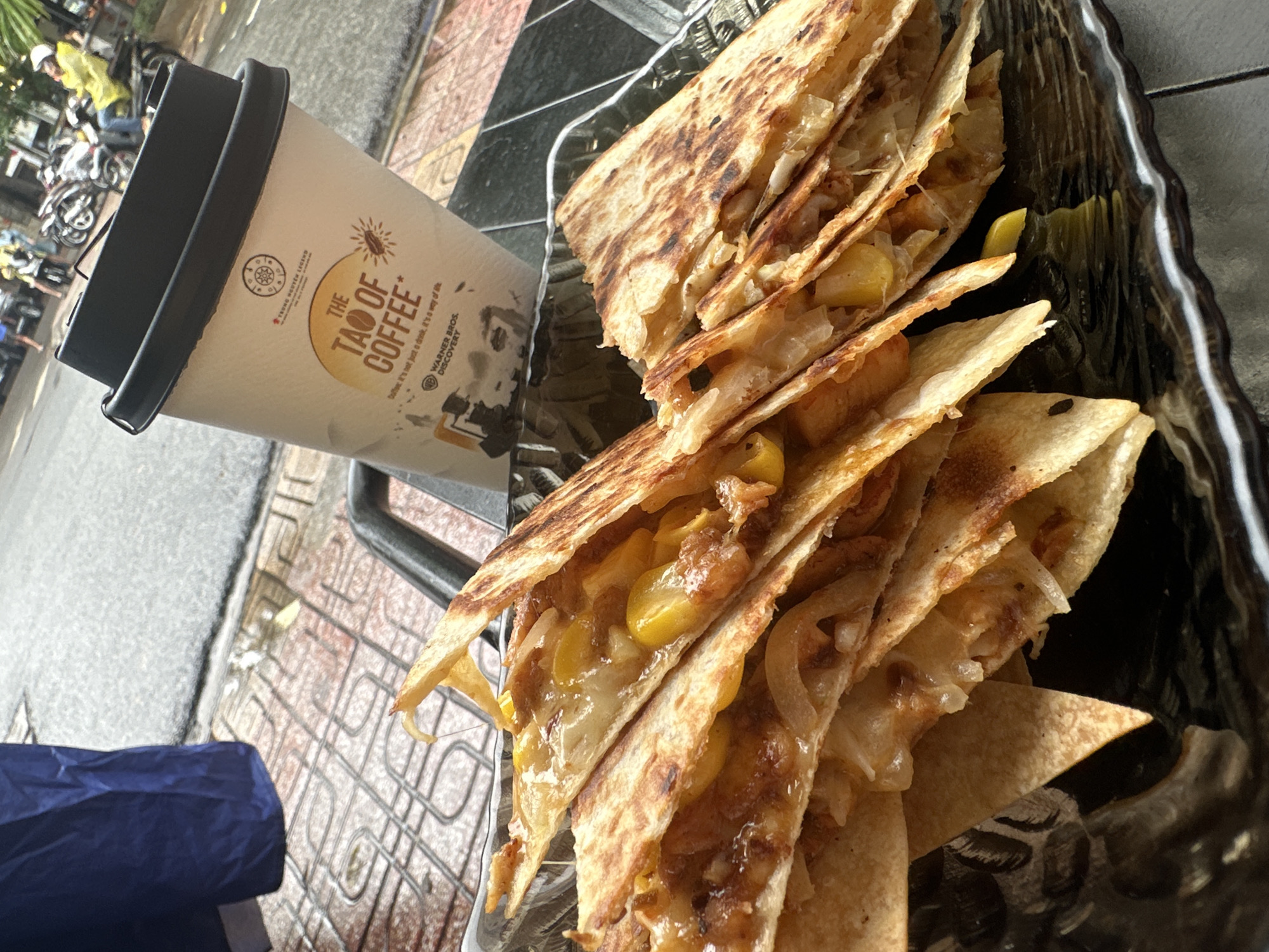 The image shows a partially obscured cup of "Tao of Coffee" from Trung Nguyen Legend sitting near a plate of quesadillas. The scene appears to be outdoors, likely in a cafe or restaurant setting. The quesadillas are arranged in a black tray, with visible fillings of what appears to be cheese, corn, and onions. In the background, there are parked motorbikes and a glimpse of street activity, suggesting the location is in an urban environment. A blue bag is partially visible in the bottom left corner. Based on the visual elements, the scene is in the Nha Trang Ward of Vietnam.
