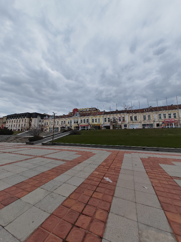 The image depicts a wide-angle view of a public square and adjacent urban landscape in Shumen, Bulgaria, under an overcast sky.
In the foreground, a paved square features a geometric pattern of alternating light gray and reddish-brown square tiles. A small, translucent white plastic bag is present on the reddish-brown tiles in the lower right quadrant.
The midground is dominated by a strip of manicured green lawn, running horizontally across the scene. To the left, a multi-tiered set of wide concrete steps ascends diagonally, bordered by a low concrete wall. A bare deciduous tree with dark, intricate branches stands near the base of these steps. Extending vertically from behind the lawn are approximately ten tall, slender, dark poles, evenly spaced, possibly flagpoles or decorative light standards.
In the background, a row of multi-story buildings is visible. To the far left, a large, ornate building with a dark mansard roof, light-colored facades, and numerous windows and visible air conditioning units stands prominently at the top of the steps. Adjacent to this, and continuing across the mid-right, is a long, continuous block of two-to-three-story structures with light yellow or cream-colored facades, showing signs of weathering and disrepair. These buildings feature multiple windows on upper floors and various ground-level storefronts with Cyrillic signage, including "БОНЖУР Д ИЕЛ" (Bonjour D'iel), "ШУМЕН-2" (Shumen-2), "ТЕЛЕНОР" (Telenor), "ОПТИКЛАЗ" (OptiClas), and "КОЗМЕТИКА" (Kosmetika). A brighter yellow building with a red roof is partially visible behind the rightmost section of this building block.
The sky occupies the upper half of the frame, completely covered by dense, textured gray clouds, indicating an overcast condition. No individuals or active movements are observed within the scene.